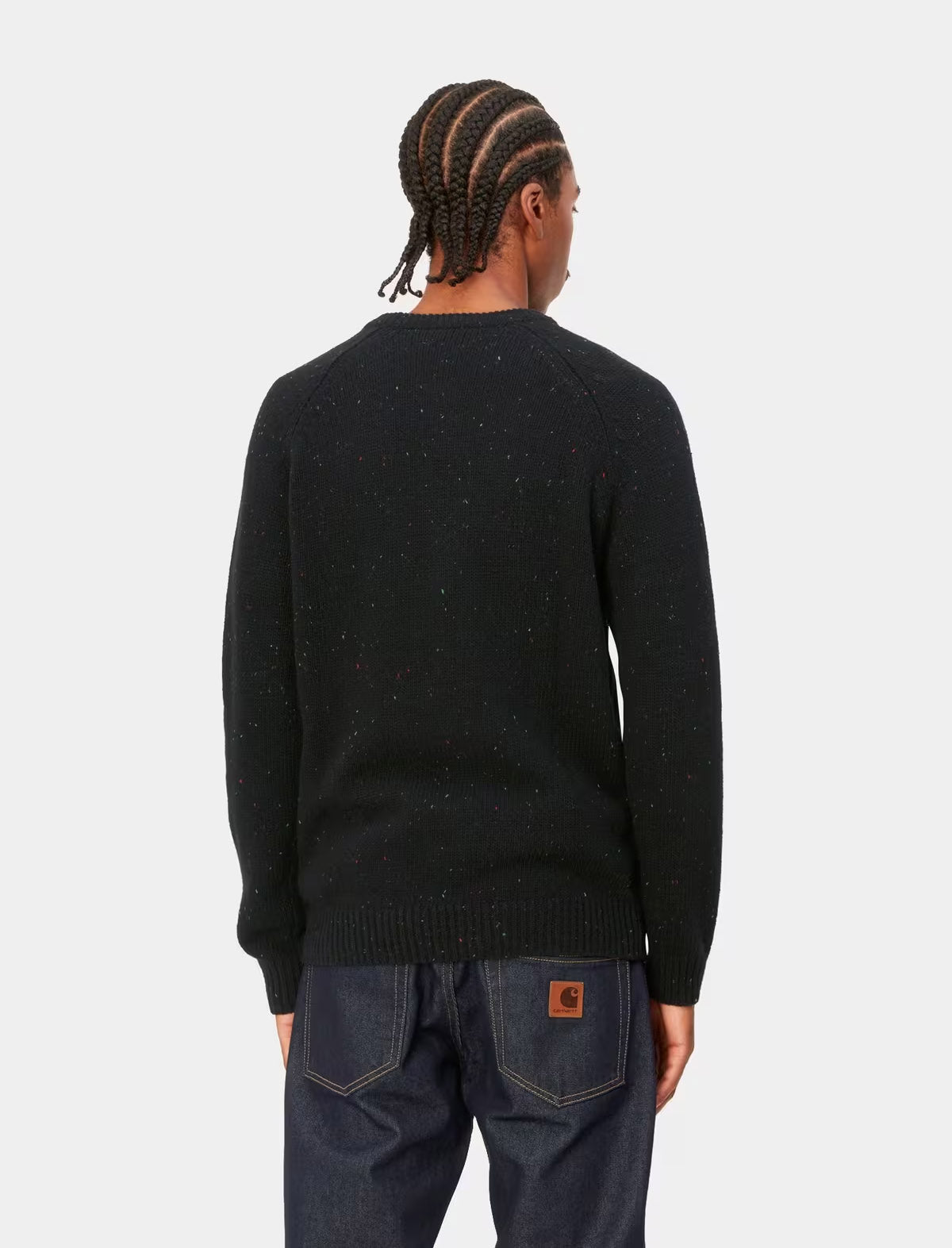 A man viewed from behind wearing a black knit sweater with subtle speckled flecks and dark blue jeans, set against a plain light gray studio background.