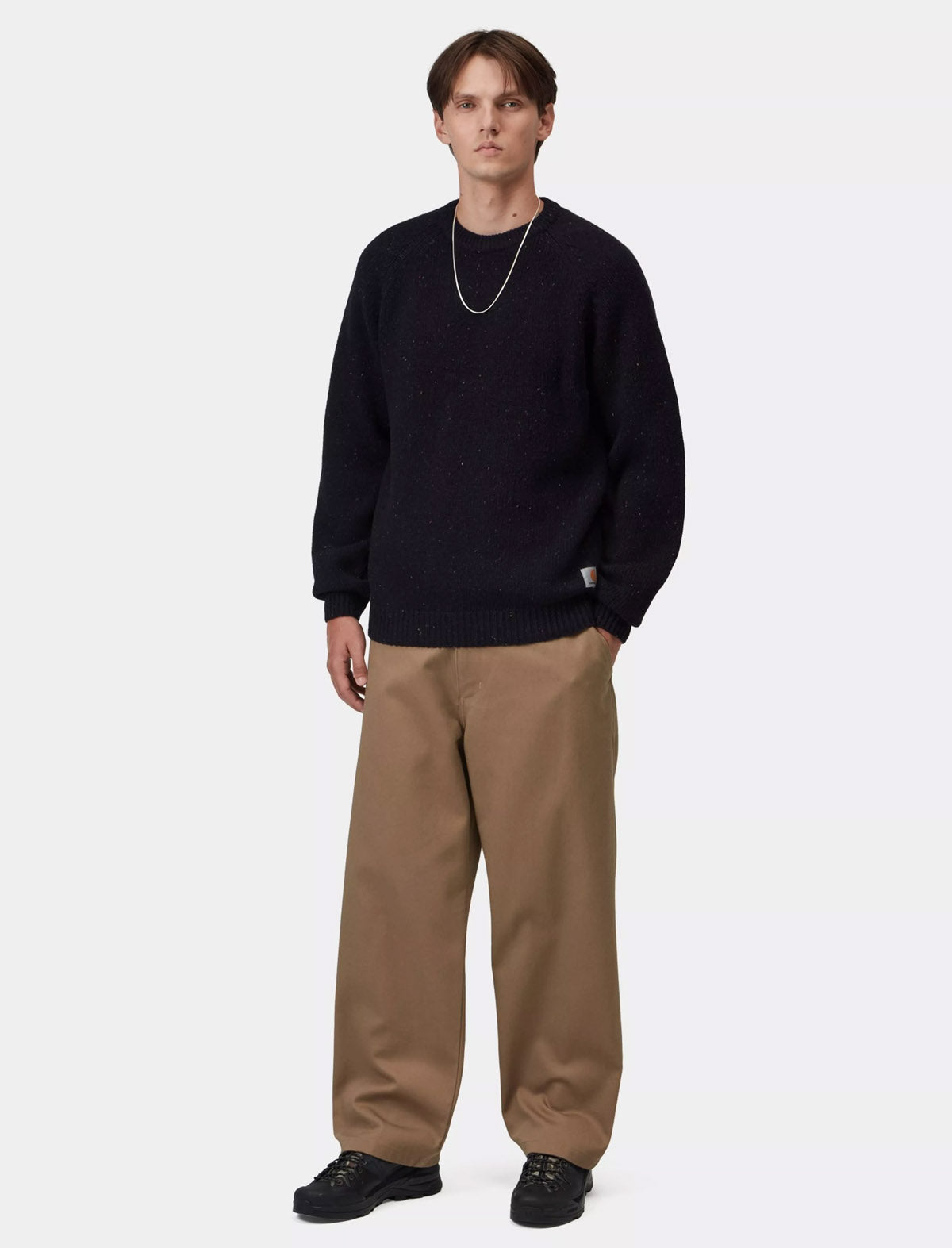 A male model wearing a dark navy knit crewneck sweater, wide-leg tan pants, and black sneakers, standing against a plain light gray studio background.