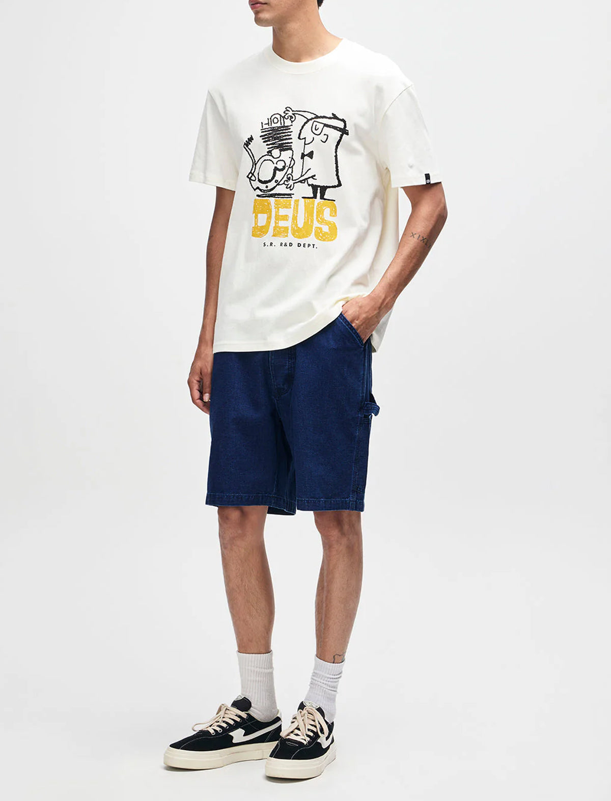 Person wearing a white t-shirt with a graphic design and text, blue shorts, and black and white sneakers on a white background.