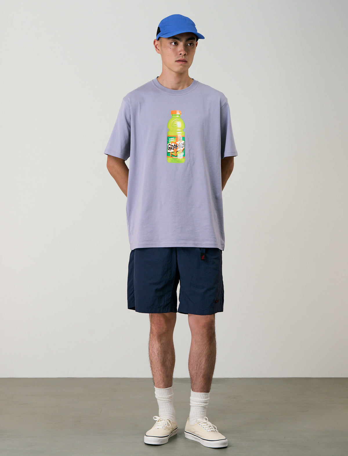 Person wearing a light purple t-shirt with a bottle graphic and navy shorts on a plain background