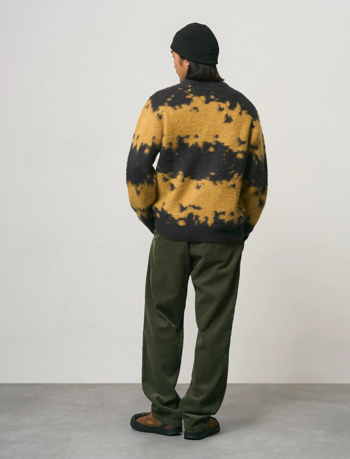 A person shown from the back wearing a fuzzy knit sweater with a black and mustard yellow tie-dye pattern, olive green pants, a black beanie, and brown shoes, standing against a plain light gray studio wall.