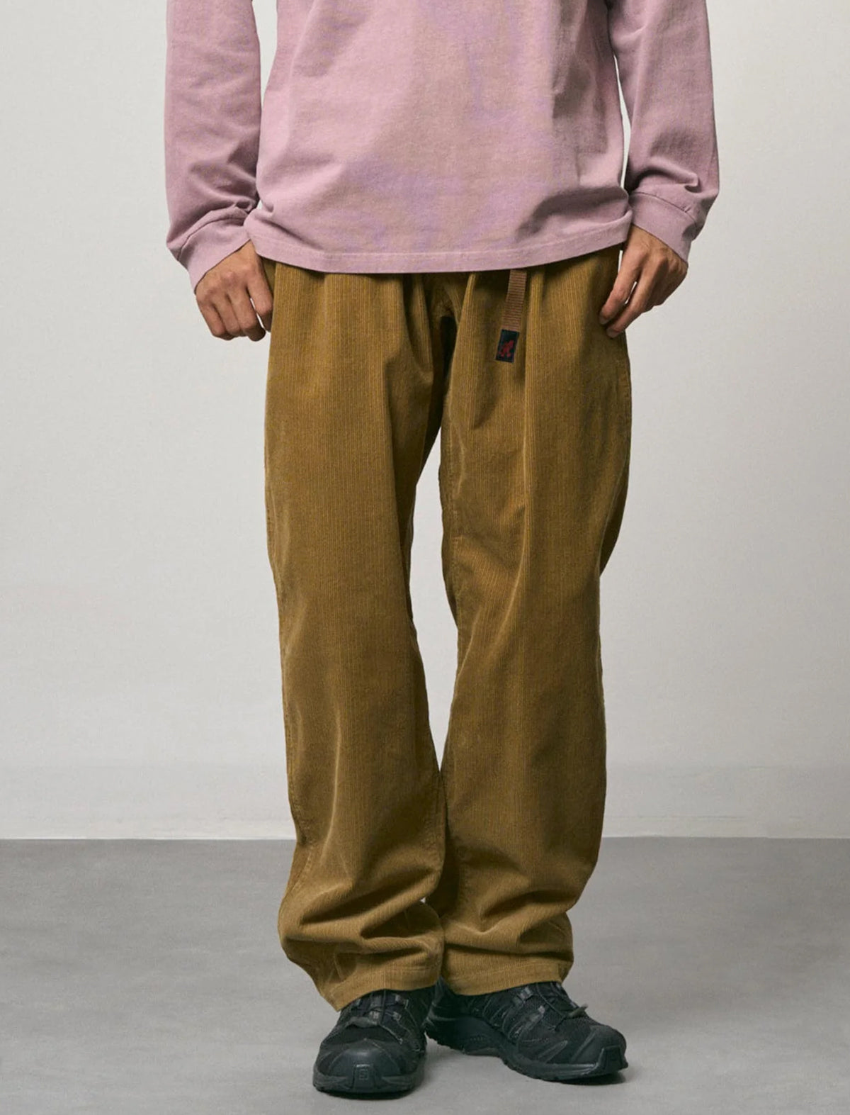 A person wearing loose brown corduroy pants with a small black brand patch on the left thigh, paired with a dusty pink long-sleeve shirt and black sneakers, standing against a plain light gray wall.
