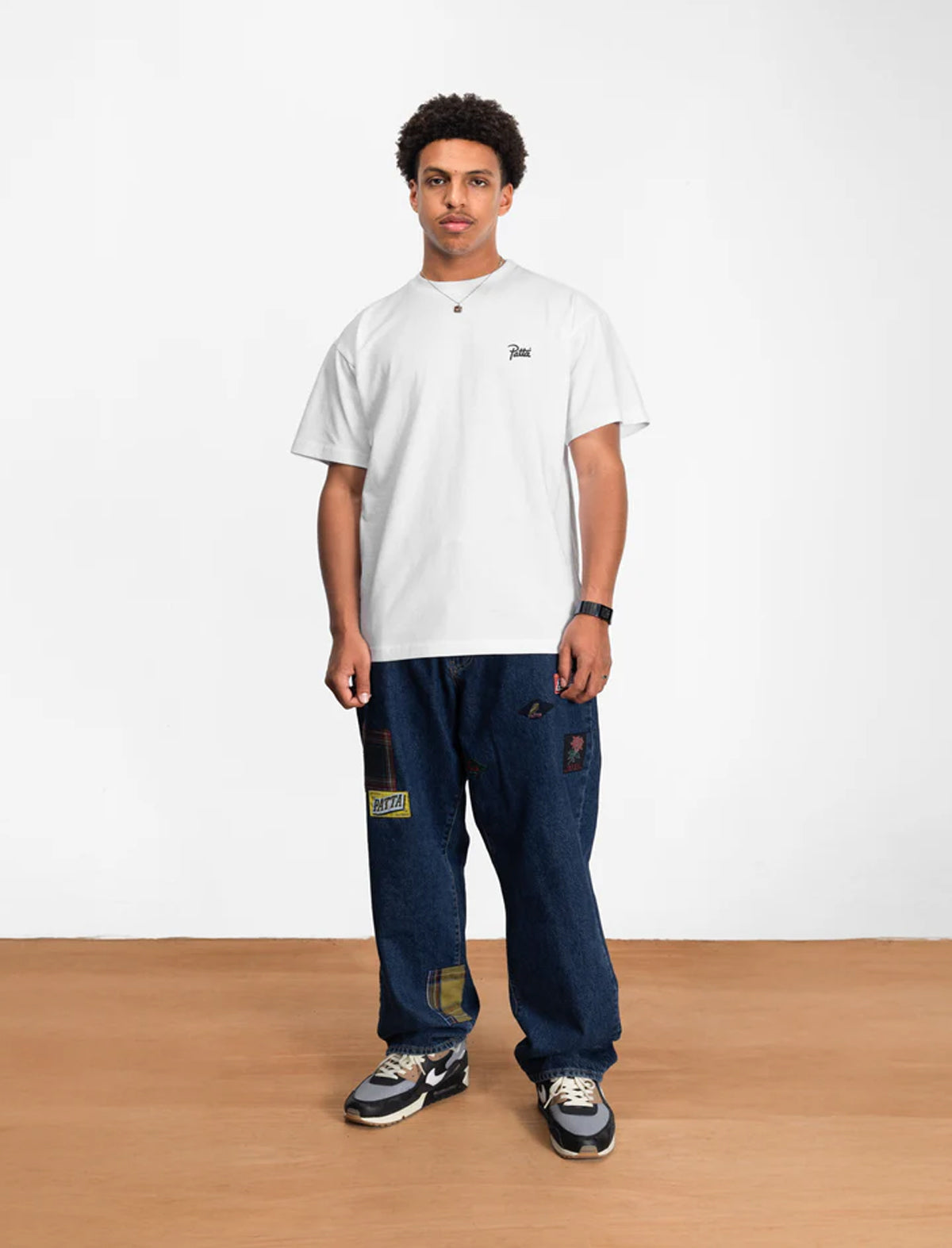 Person wearing a white t-shirt and blue jeans standing against a white wall.