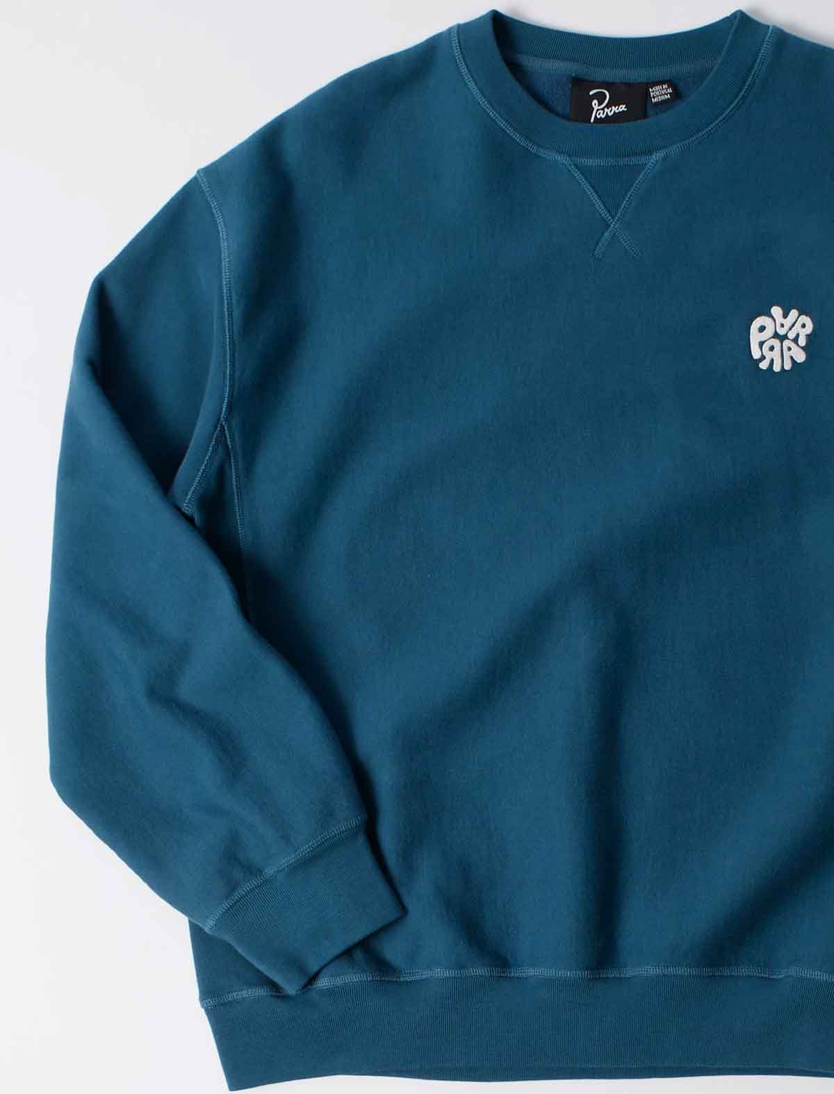 A teal blue crewneck sweatshirt with a small white embroidered graphic on the left chest, displayed flat against a clean white background.