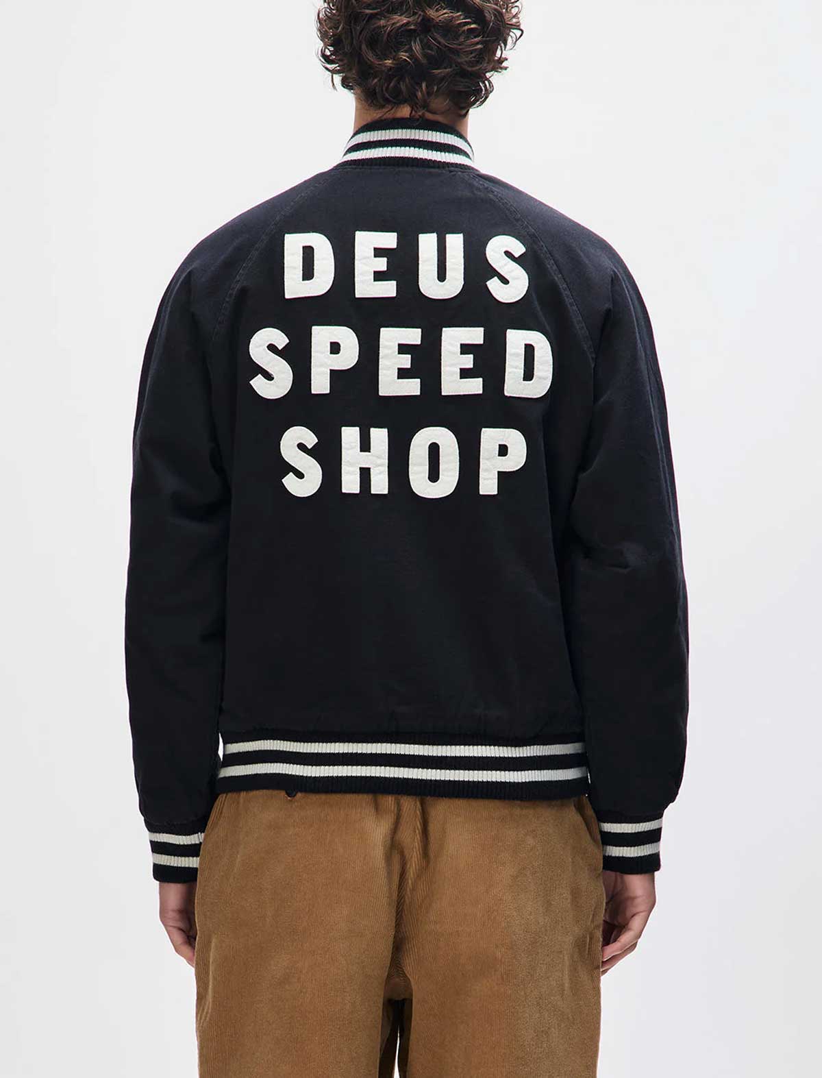 Person wearing a black jacket with 'DEUS SPEED SHOP' text on the back against a white background