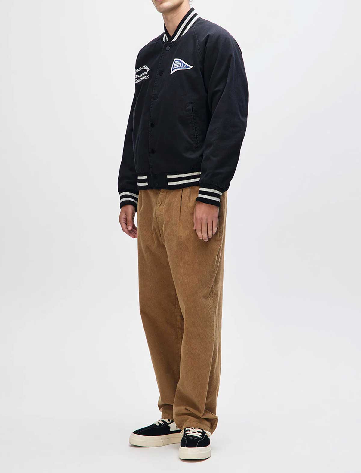 Person wearing a black varsity jacket with white stripes and brown pants on a white background