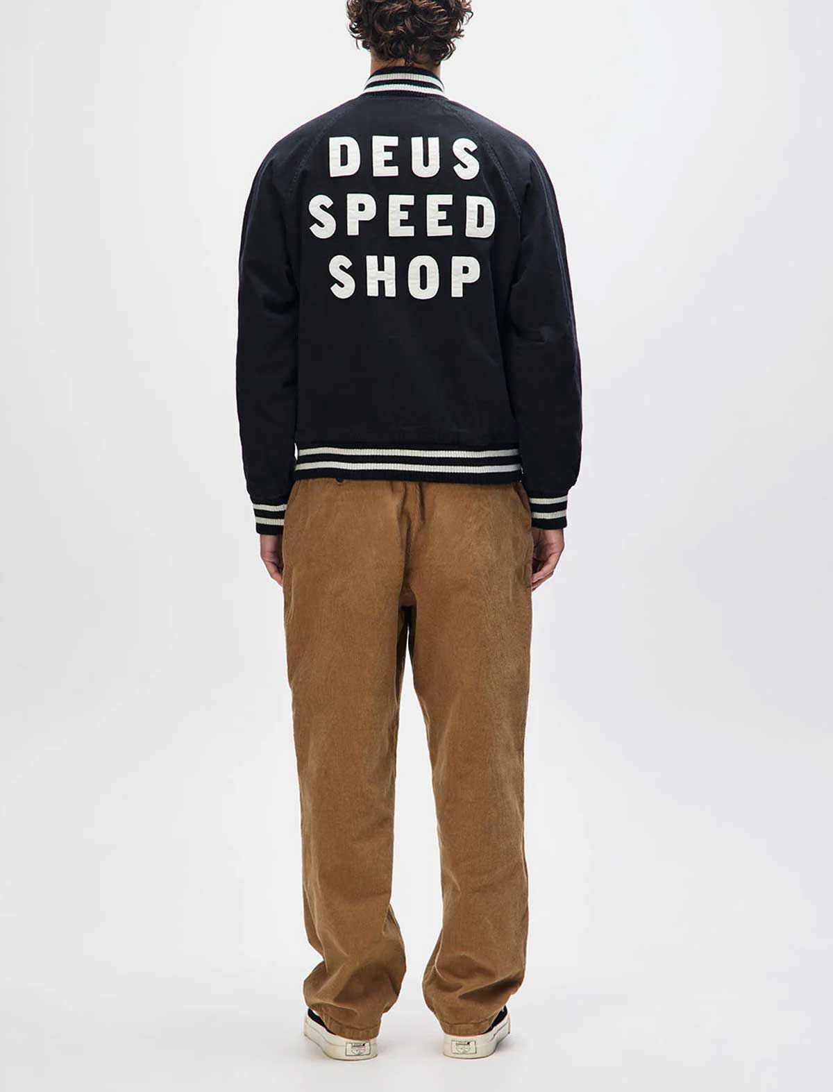 Person wearing a black jacket with 'DEUS SPEED SHOP' text on the back, standing against a white background.