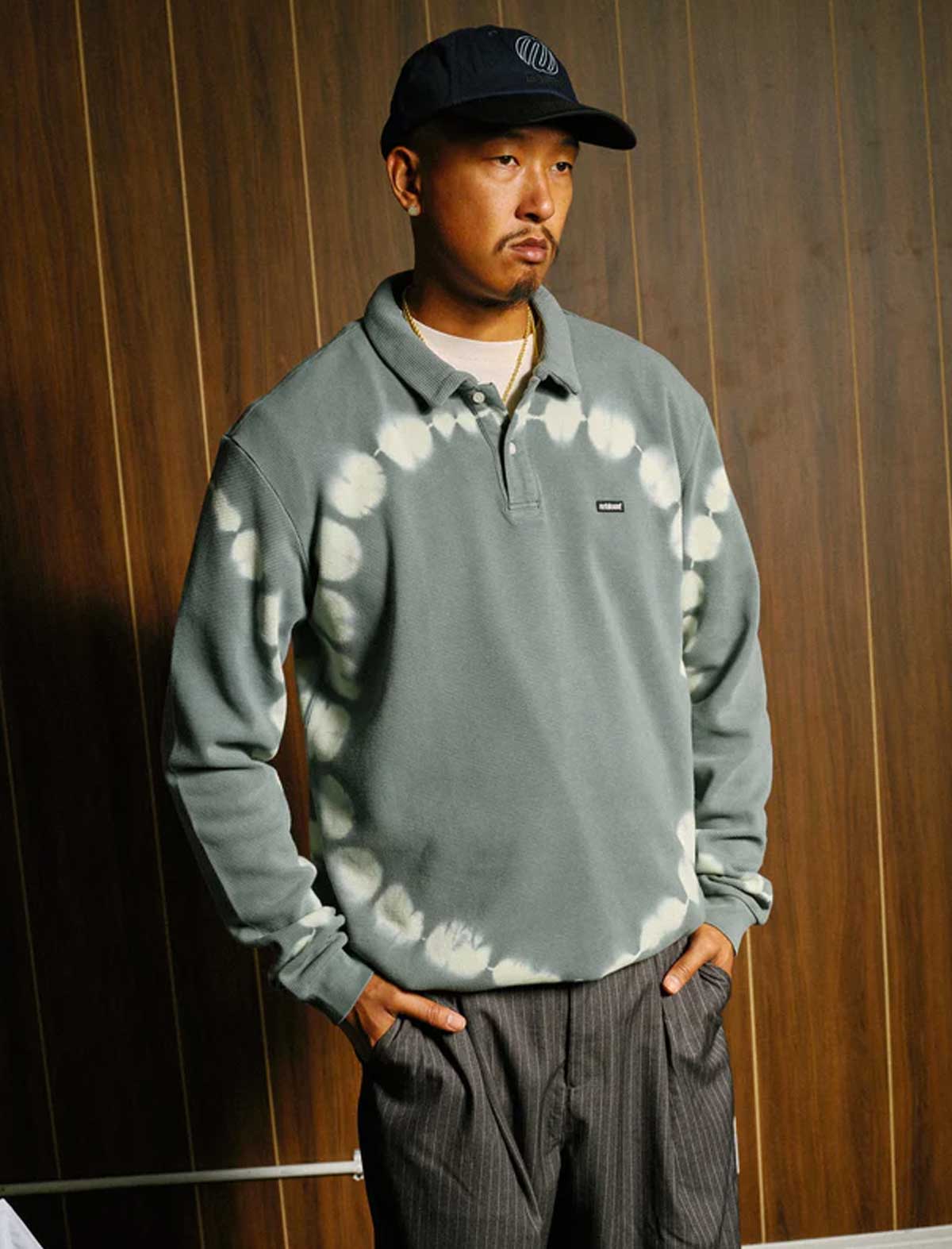 Person wearing a Metalwood gray tie-dye sweater and cap against a wooden panel background