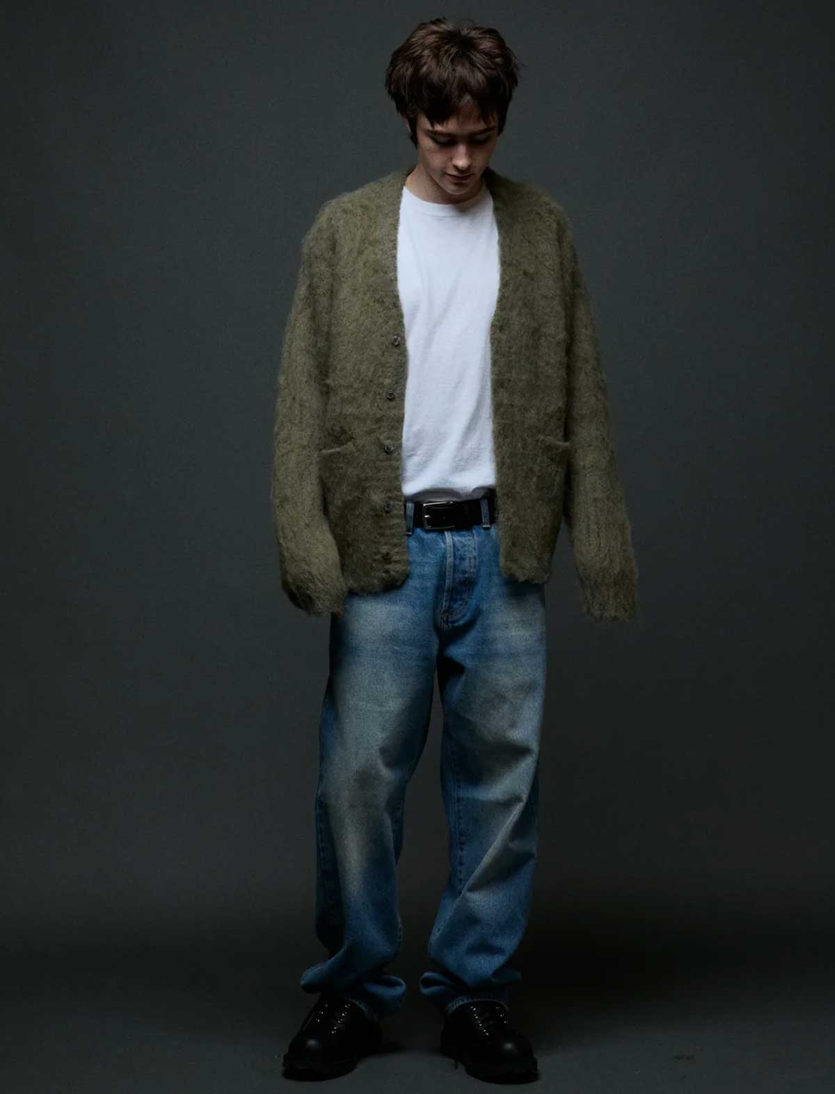 A man wearing a fuzzy olive green cardigan over a white t-shirt, blue jeans, and black shoes, standing against a dark gray studio background.