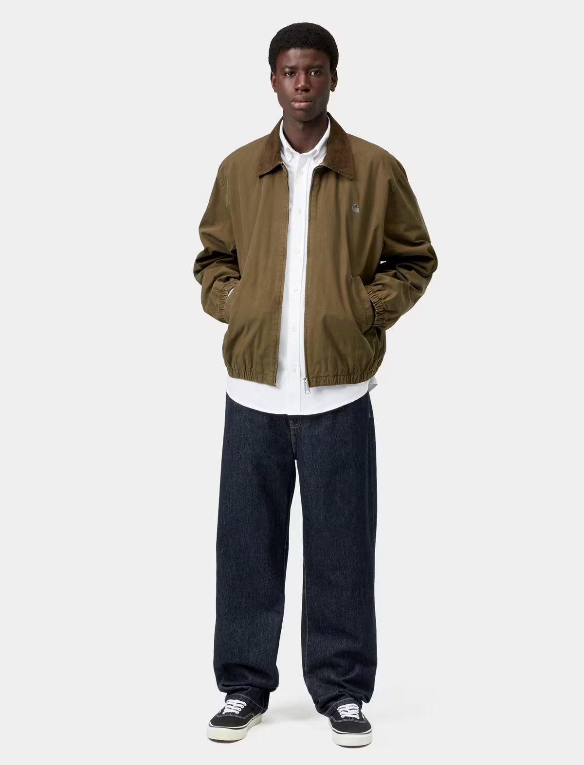 A model stands against a light gray studio background wearing an olive green jacket over a white collared shirt, dark blue jeans, and black sneakers.