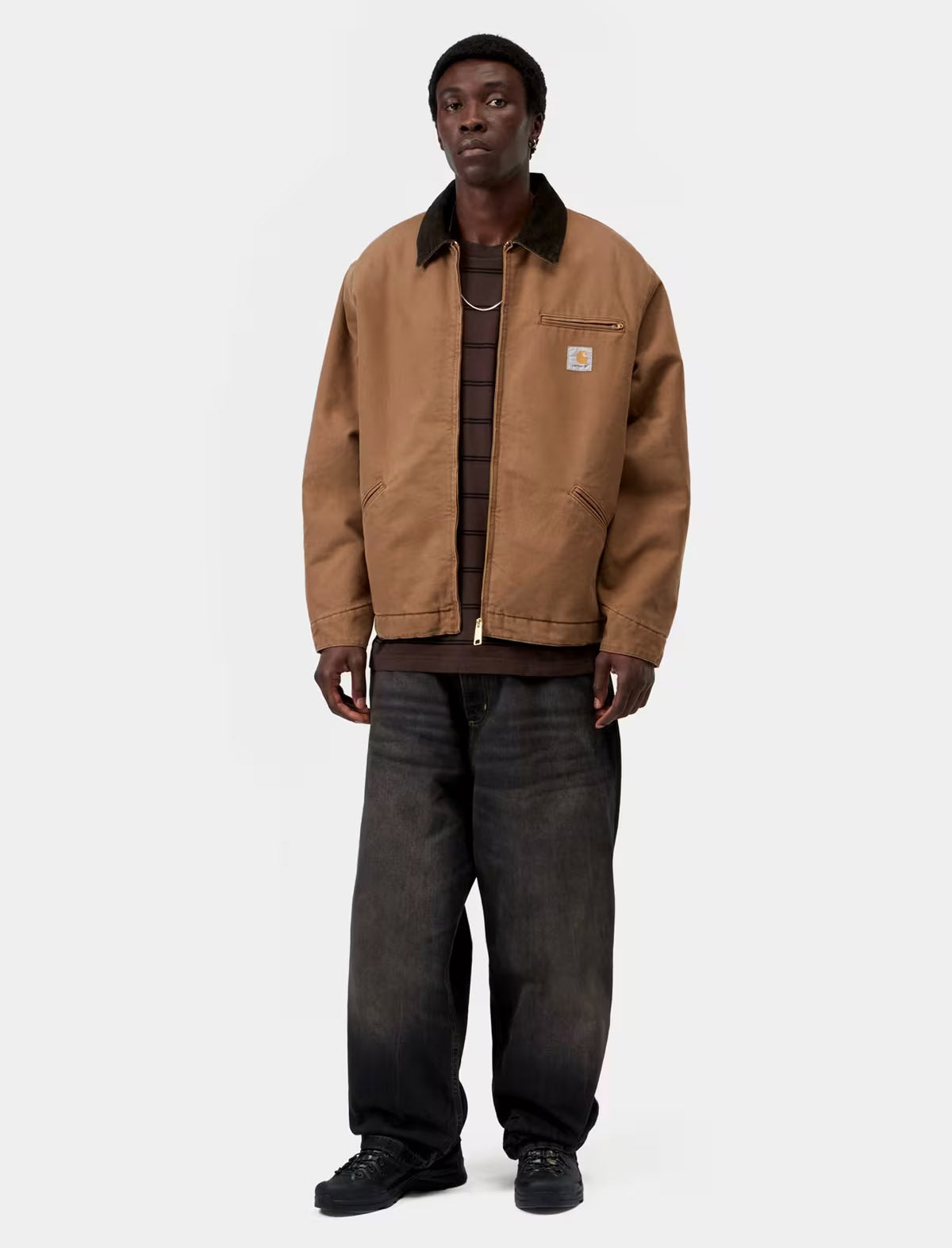 A model wearing a brown Carhartt jacket with a dark brown corduroy collar, layered over a dark brown striped shirt and dark gray jeans, standing against a light gray background.