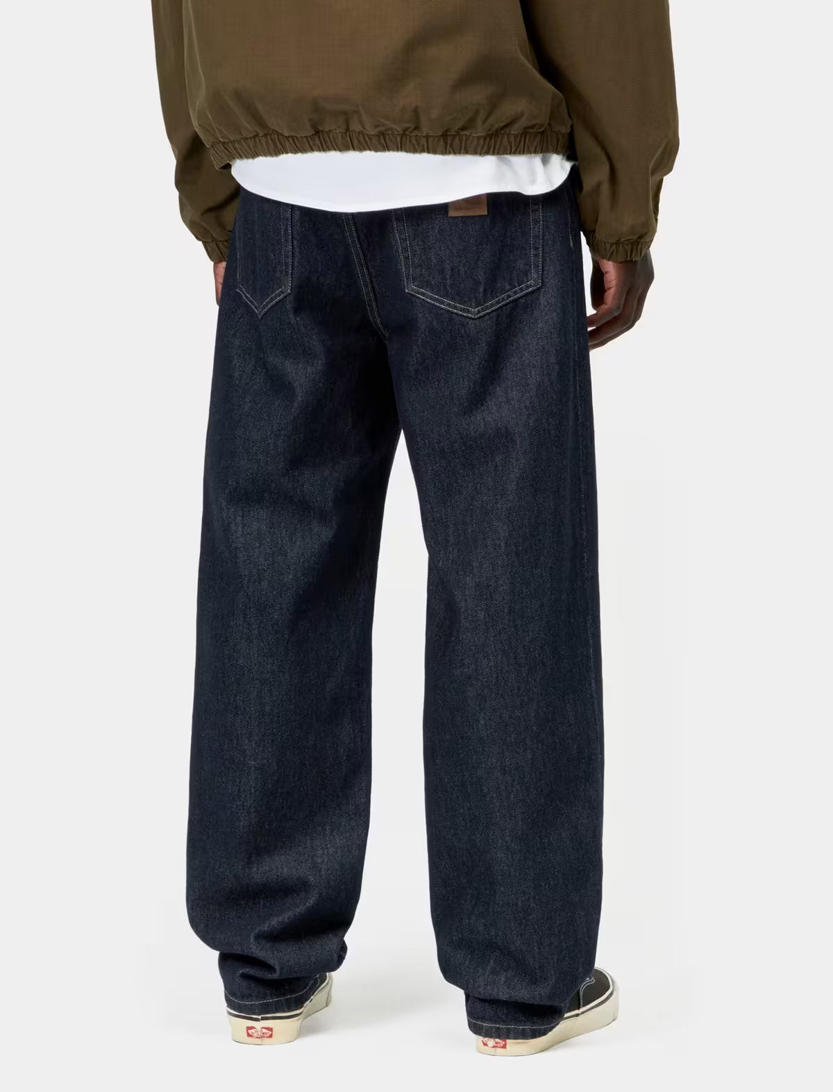 Rear view of a person wearing dark indigo wide-leg jeans, a white t-shirt, and an olive green jacket, standing against a light gray studio background.