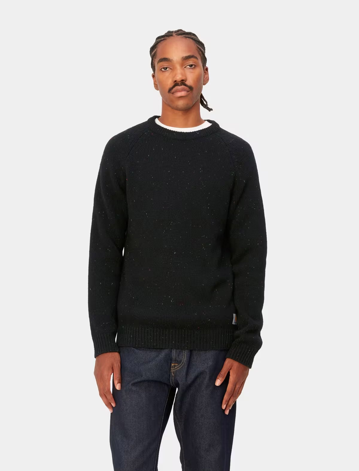 A man wearing a black knit crewneck sweater with subtle speckled texture, paired with dark blue jeans, standing against a light gray background.