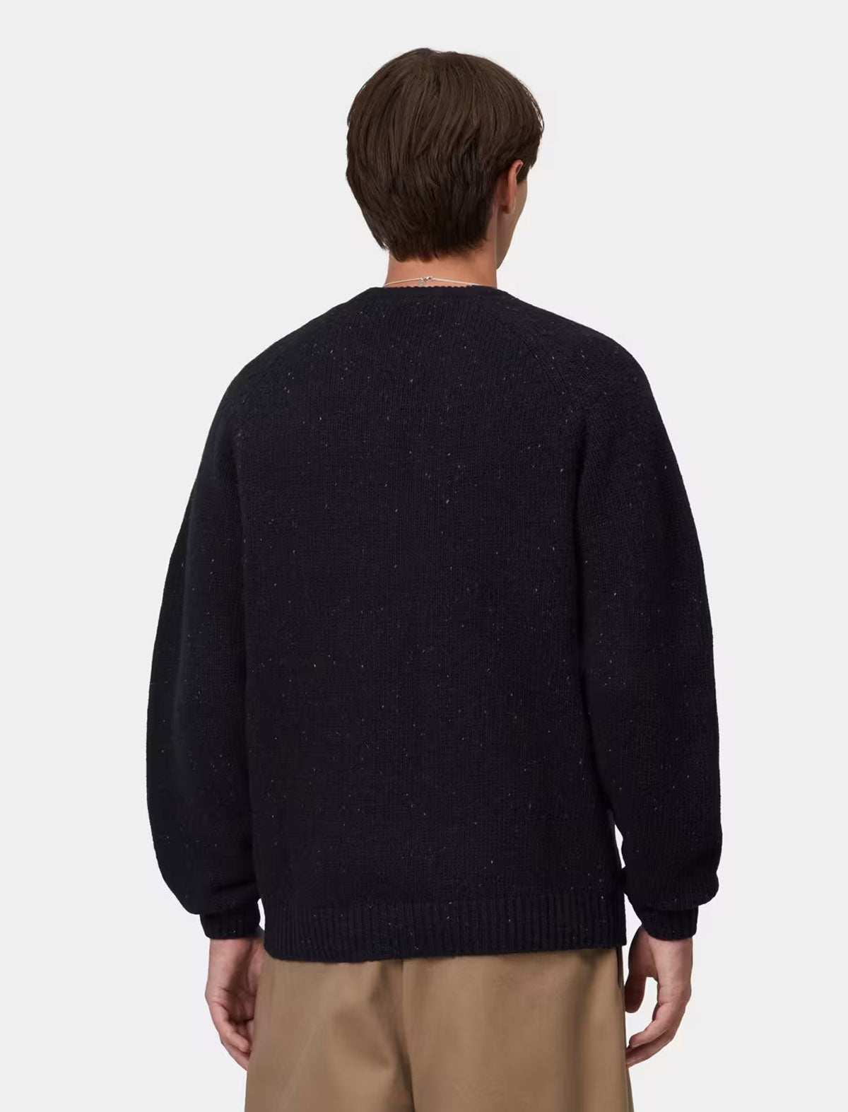 A man viewed from behind wearing a dark navy knit sweater with subtle flecks and beige trousers, set against a plain light gray background.