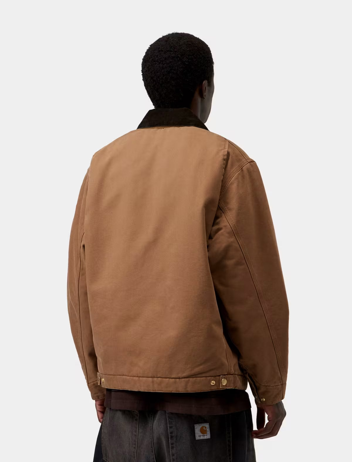 A person shown from the back wearing a tan Carhartt jacket with a dark collar, paired with dark jeans, against a light gray studio background.