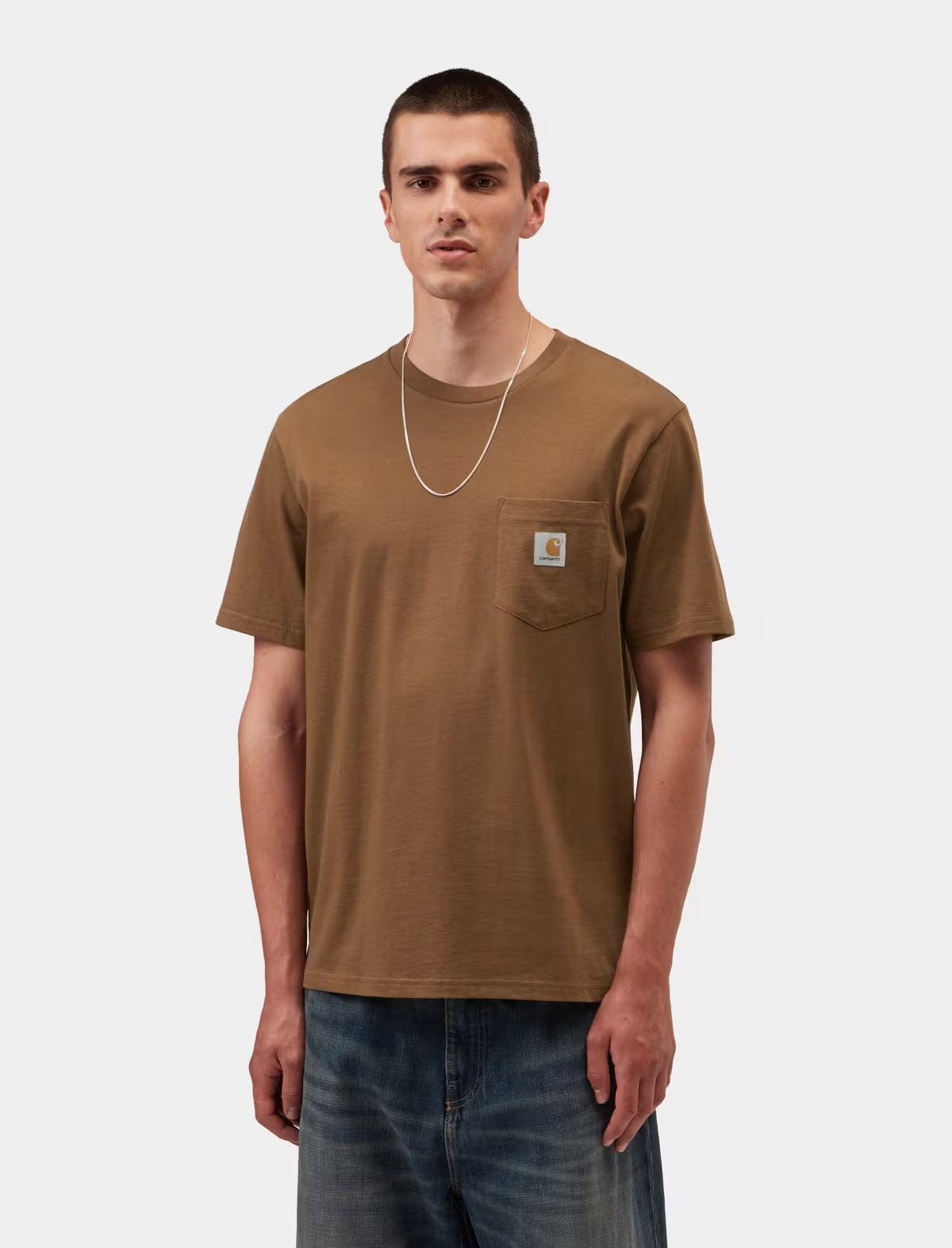 A man wearing a brown short-sleeve pocket t-shirt with a small Carhartt logo patch on the chest pocket, paired with blue jeans and a silver chain necklace, against a light gray background.
