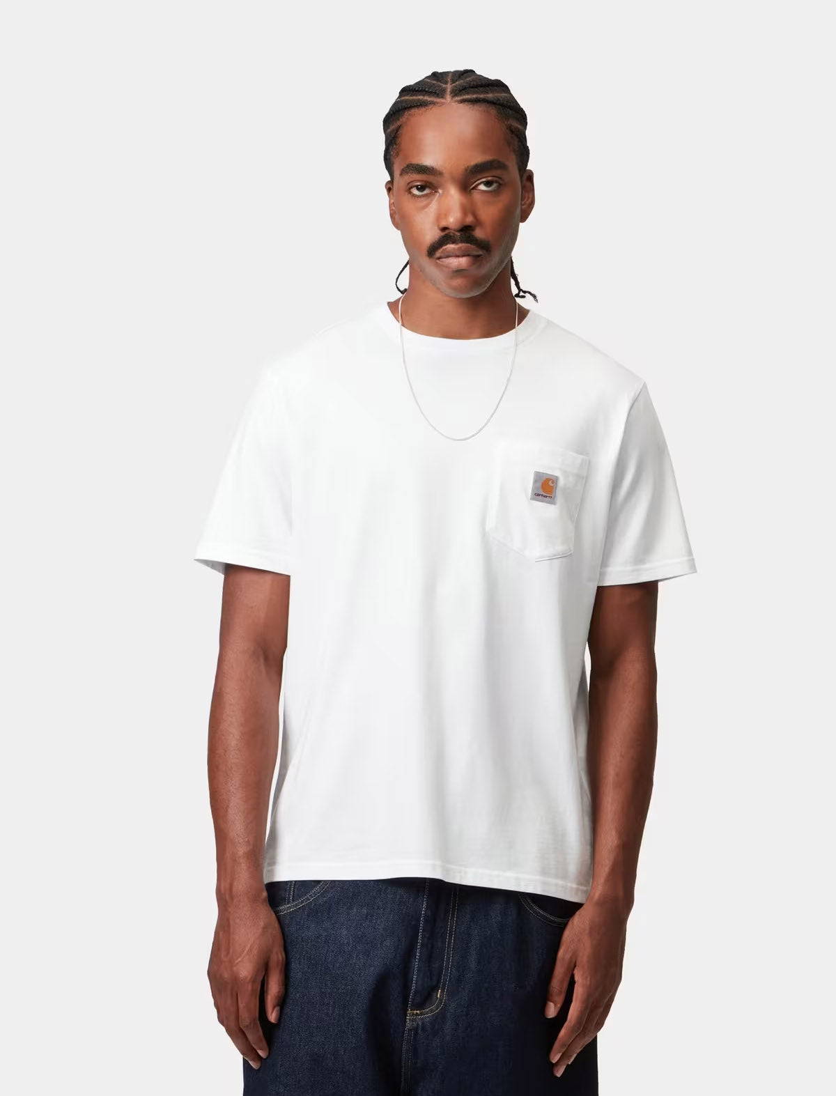A man wearing a white short-sleeve pocket t-shirt by Carhartt, paired with dark blue jeans, standing against a light gray studio background.