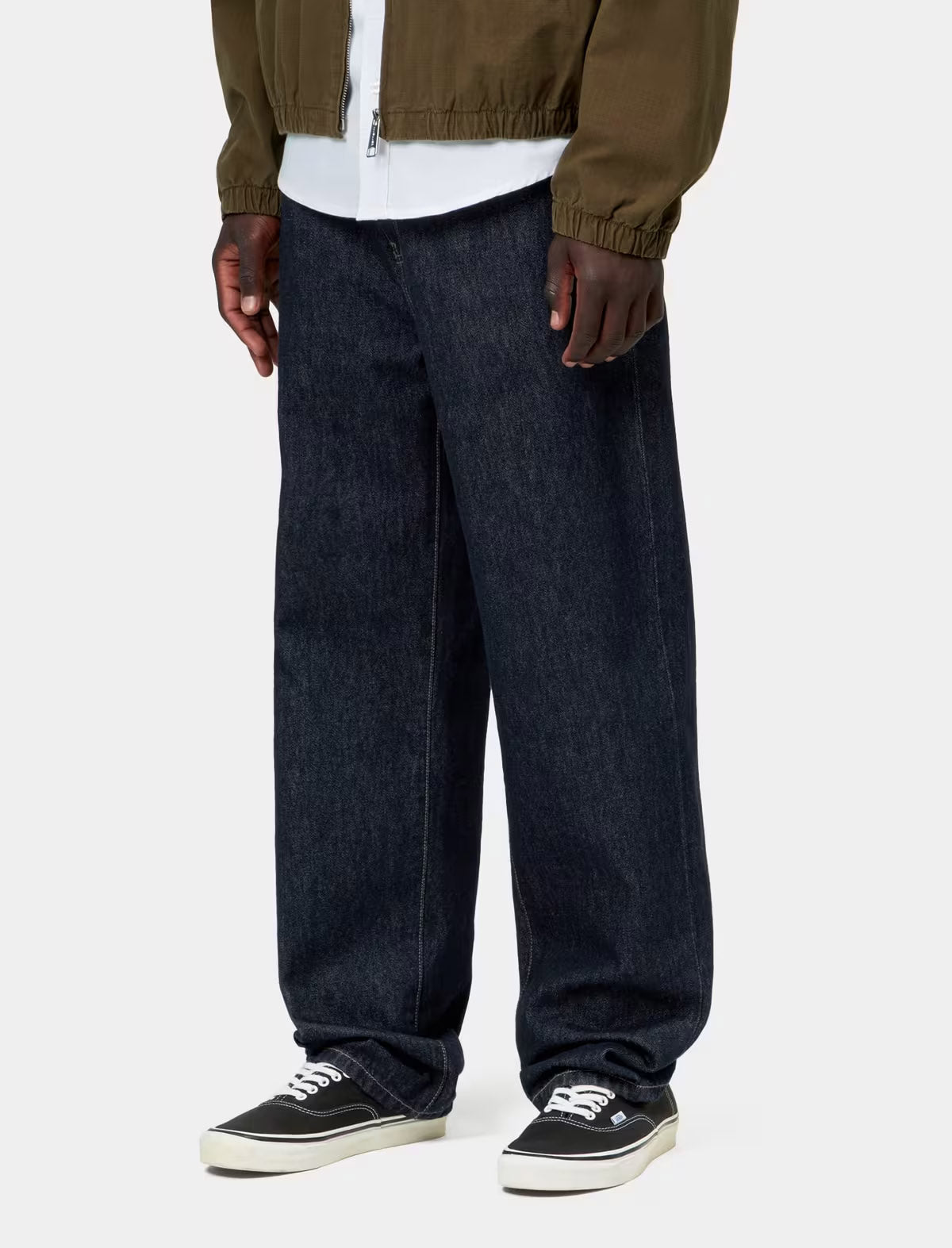 A person wearing loose dark blue denim jeans, a white t-shirt, an olive green jacket, and black low-top sneakers, shown from the waist down against a light gray studio background.