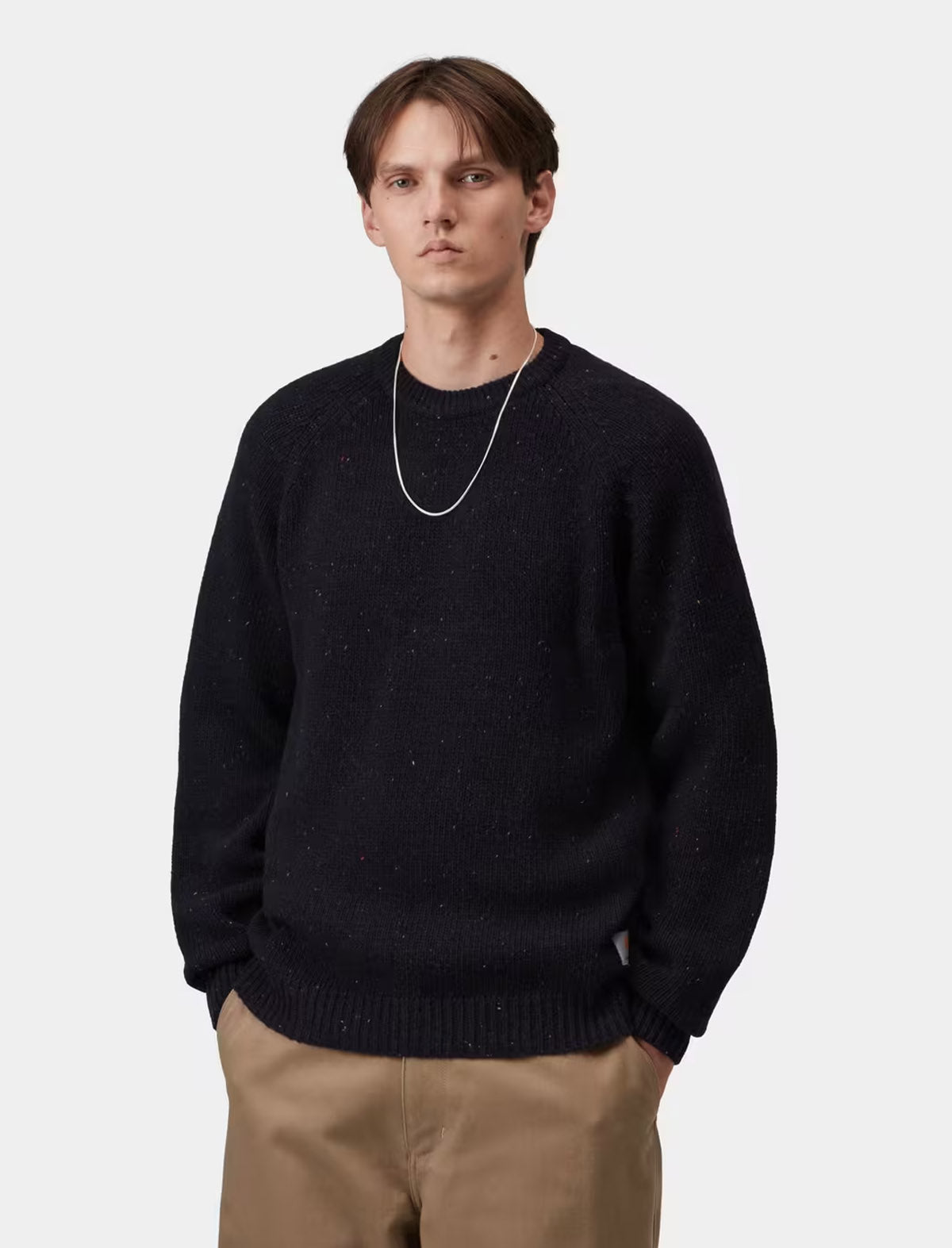 A man wearing a dark navy knit crewneck sweater with subtle speckles, paired with tan pants and a silver chain necklace, photographed against a light gray studio background.