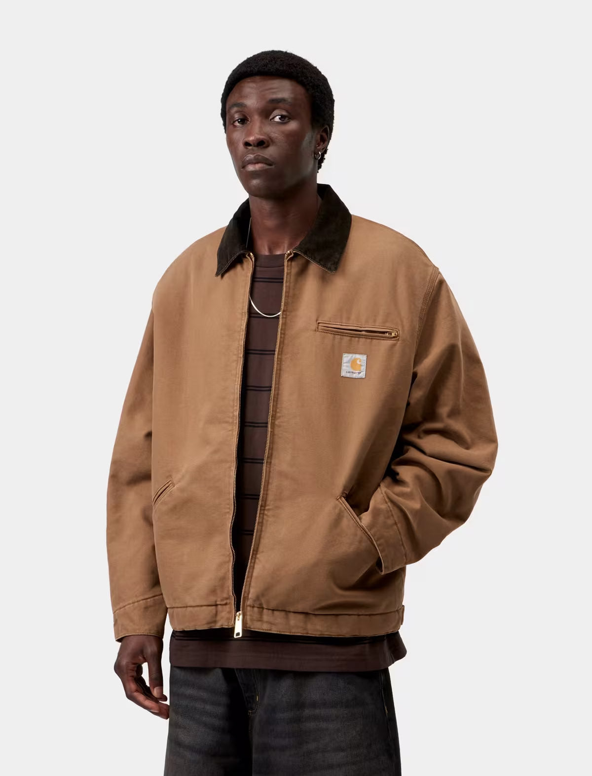 A man wearing a brown Carhartt jacket with a dark corduroy collar, layered over a dark striped shirt and dark jeans, photographed against a light gray studio background.