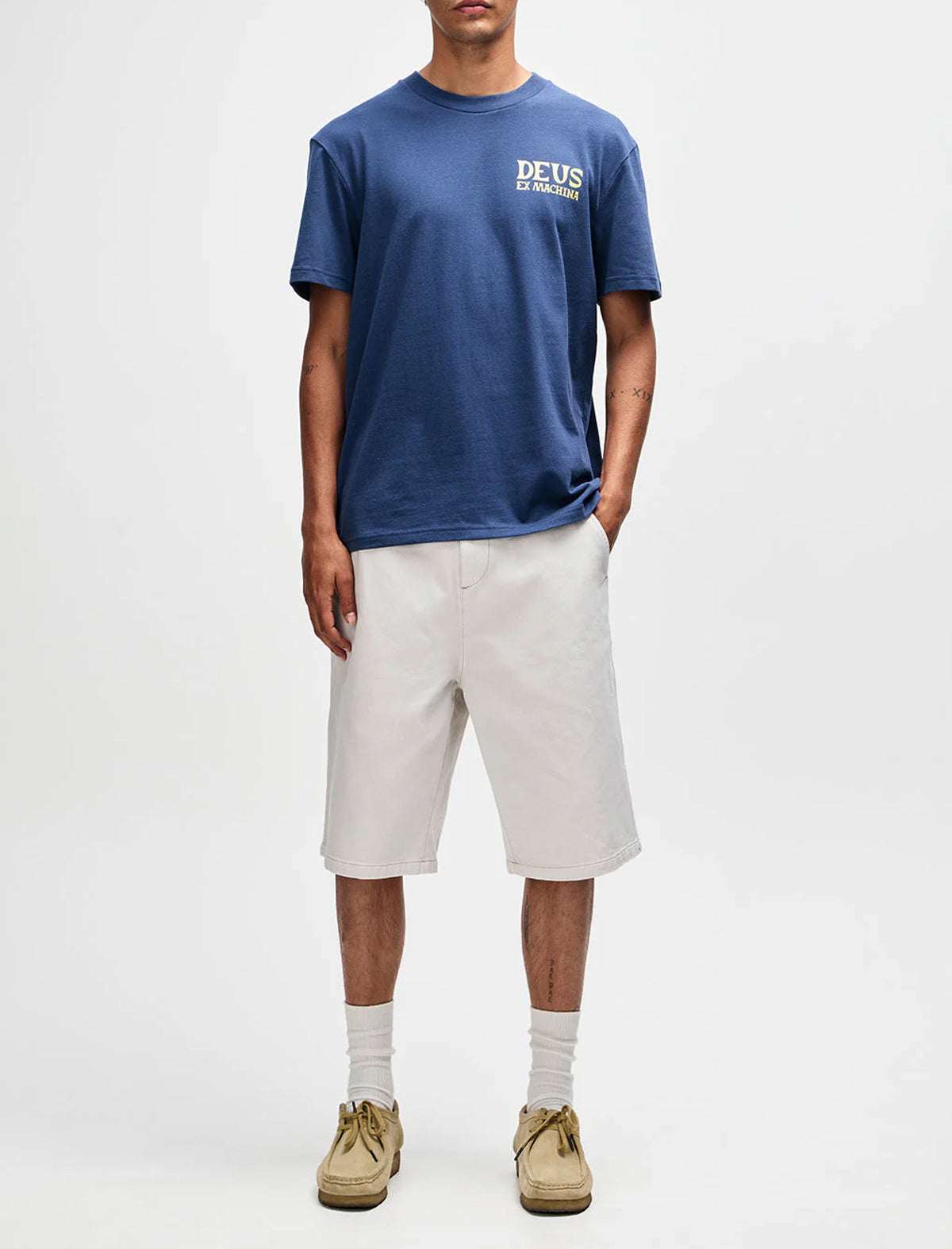 Person wearing a blue t-shirt with 'DEUS' logo and beige shorts on a white background
