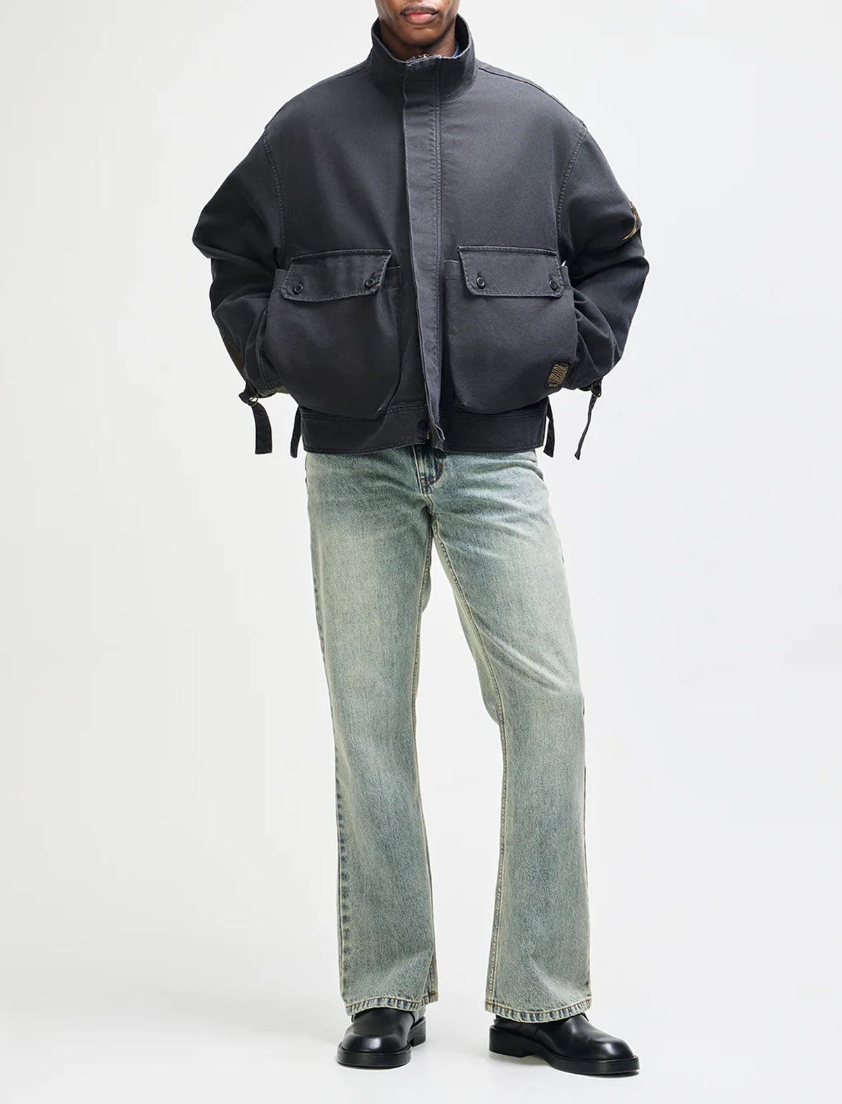 A model wearing a dark gray cropped utility jacket over a gray shirt, paired with light-wash blue jeans and black leather shoes, photographed against a solid light gray background.