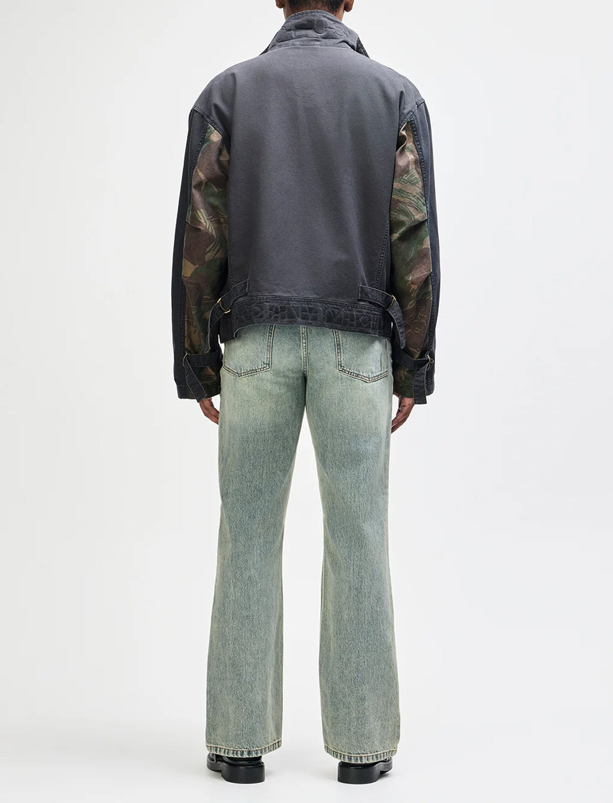 A model shown from the back wearing a dark gray jacket with camouflage-patterned sheer sleeves and light-wash blue flare jeans, against a plain white studio background.