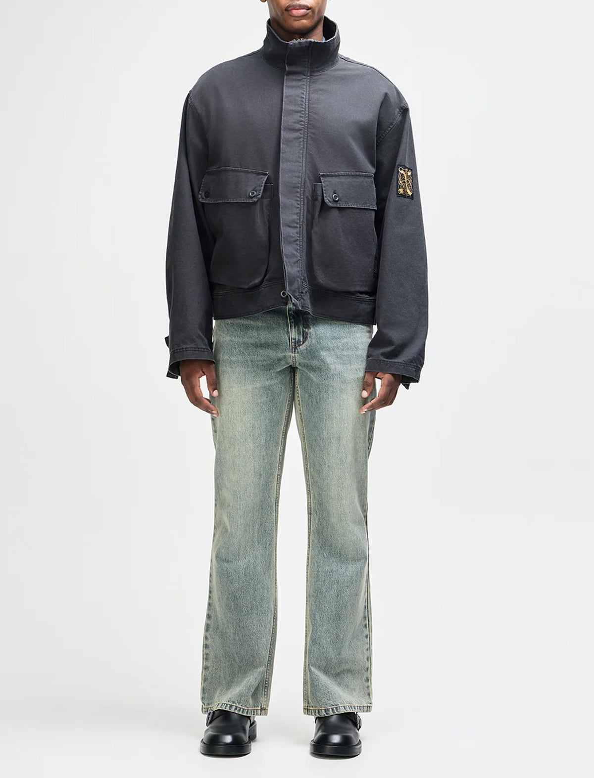 A model wearing a dark gray zip-up utility jacket with flap pockets and a Stüssy logo patch on the sleeve, paired with light-wash blue jeans and black shoes, against a plain white studio background.
