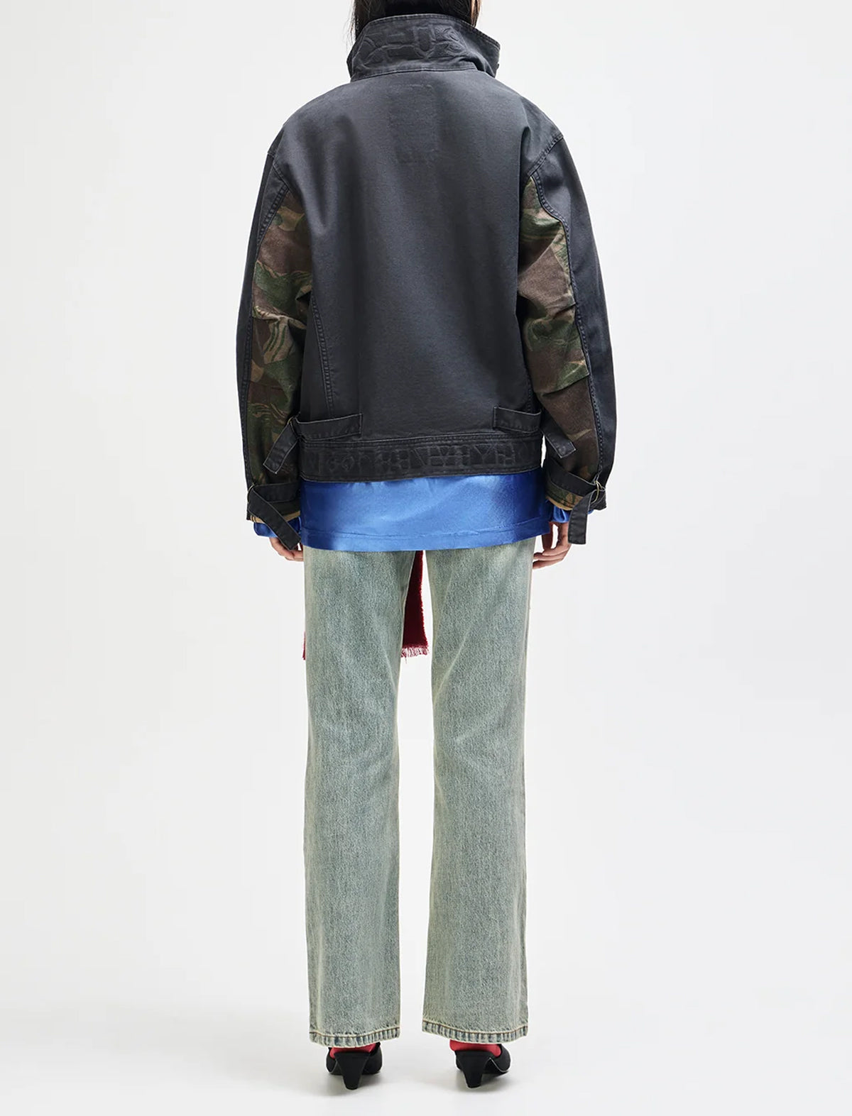A model shown from the back wearing a dark gray utility jacket with camouflage-patterned sleeves, a blue shirt underneath, light-wash wide-leg jeans, and black shoes, against a plain white studio background.