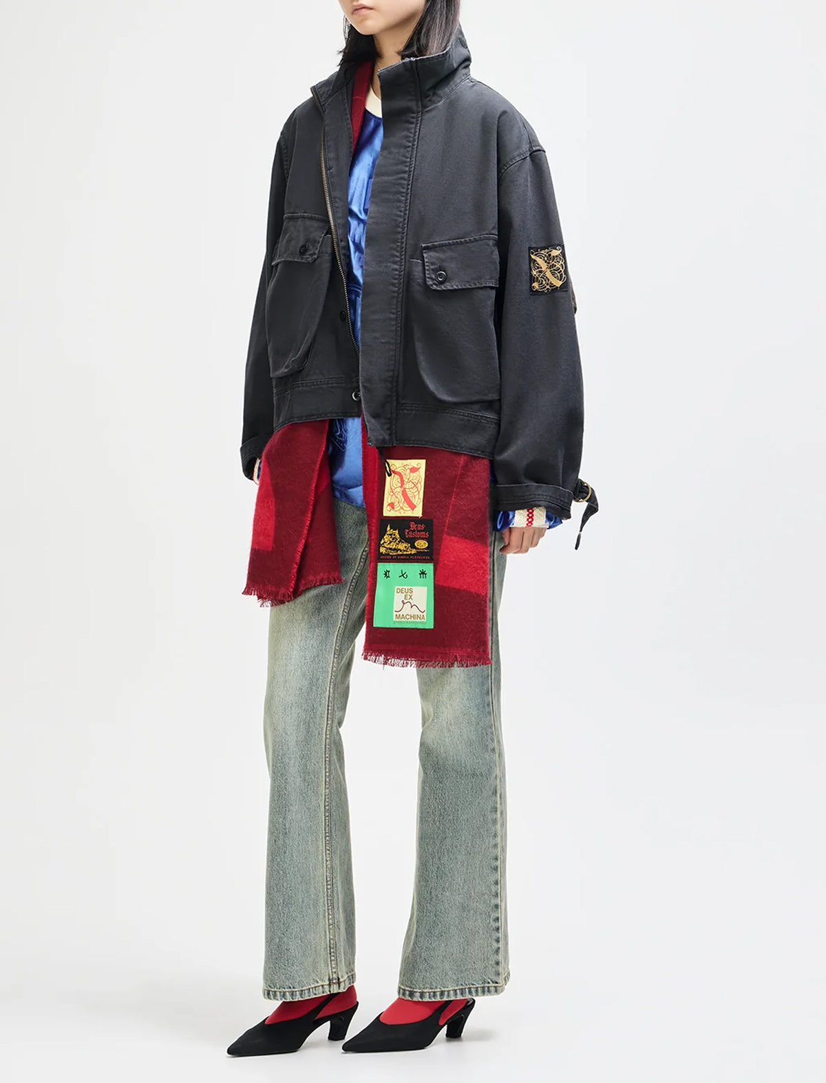 A model is shown from the neck down wearing a black utility jacket with a colorful patch on the sleeve and a red-and-black fringed patch on the front, styled with a blue shirt, light-wash flared jeans, and black pointed-toe heels against a plain white studio background.
