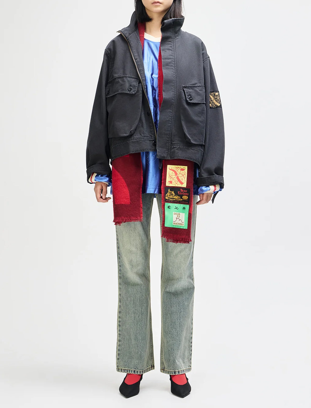 A model is shown from the neck down wearing a dark gray utility jacket with colorful patchwork details, a blue shirt, a red scarf, wide-leg light-wash jeans, and red socks against a plain white studio background.