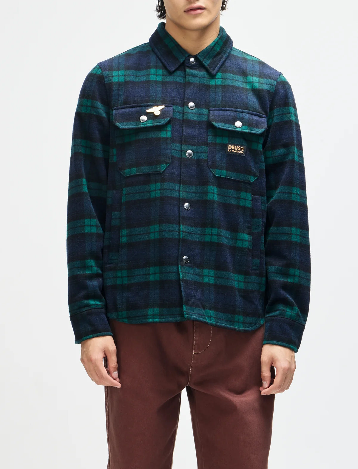 A model wearing a dark green and navy blue plaid button-up shirt jacket with a small embroidered logo on the chest pocket, paired with brown pants, against a plain white background.