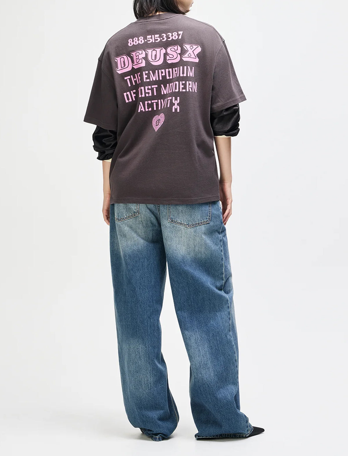 A person shown from the back wearing a dark charcoal graphic T-shirt with pink text reading 'DEUS' and additional text below, paired with wide-leg blue jeans and black shoes, against a plain light gray studio background.