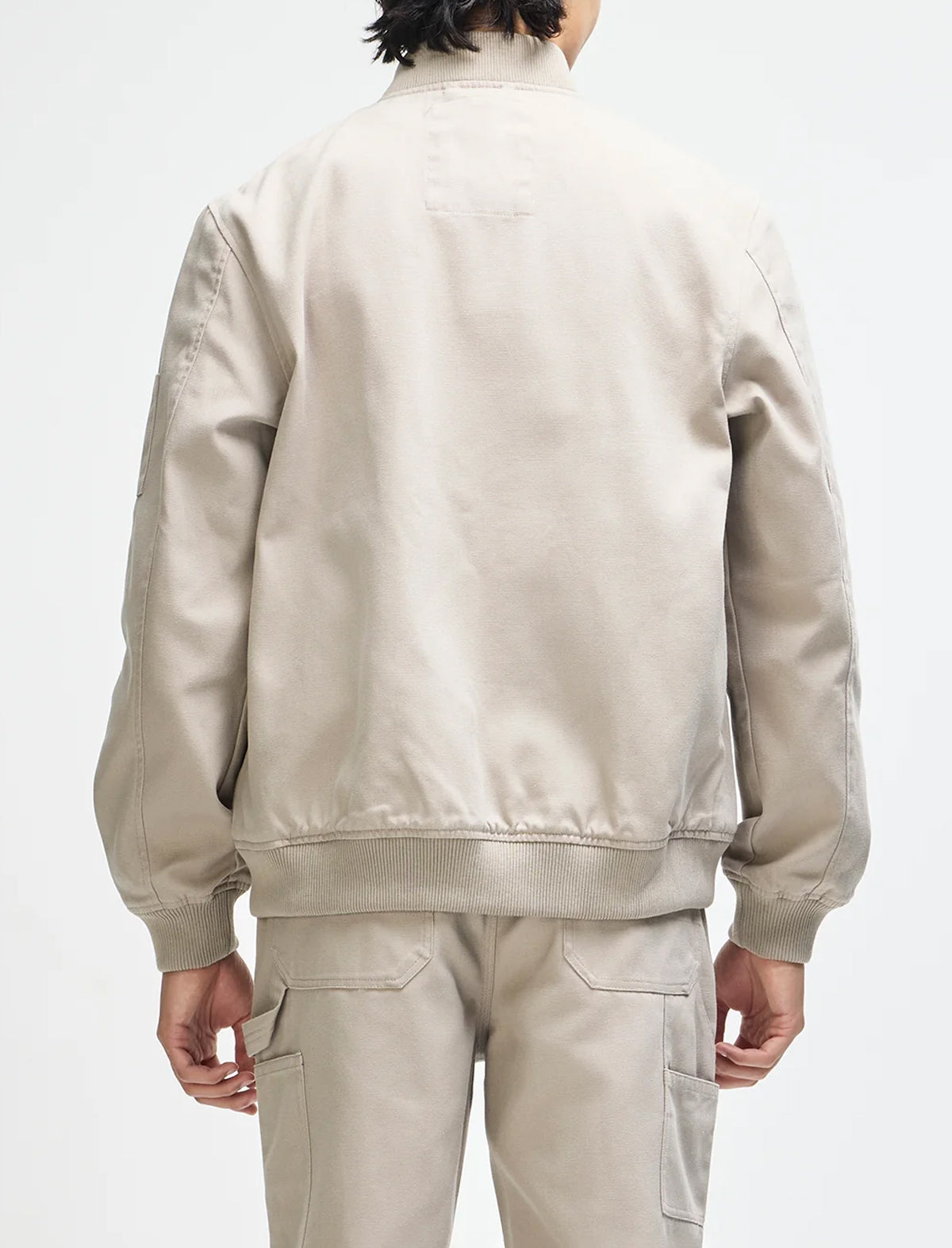 A person shown from the back wearing a light beige bomber jacket and matching beige cargo pants against a plain light gray studio background.
