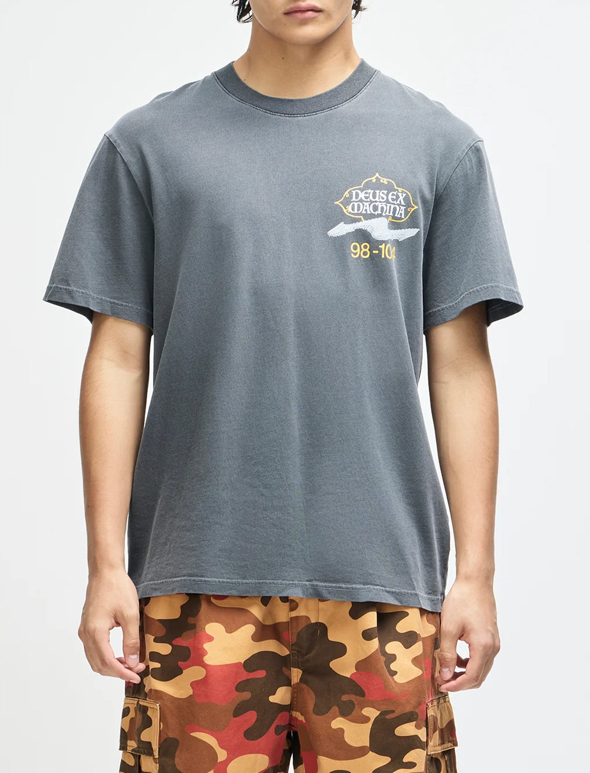 A person wearing a faded dark gray crewneck t-shirt with a small white and yellow graphic on the left chest, paired with brown, orange, and black camouflage shorts, against a plain light gray background.