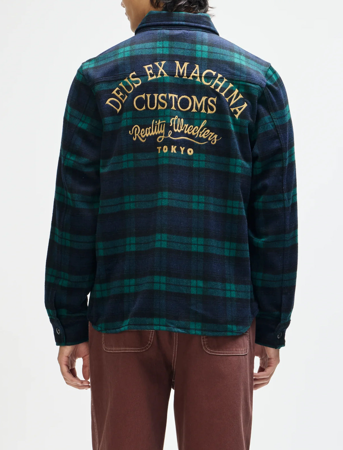 A person shown from the back wearing a long-sleeve plaid flannel-style shirt jacket with large gold text reading 'DEUS EX MACHINA CUSTOMS Reality Wreckers TOKYO' across the upper back. The garment has a dark navy and green plaid pattern.