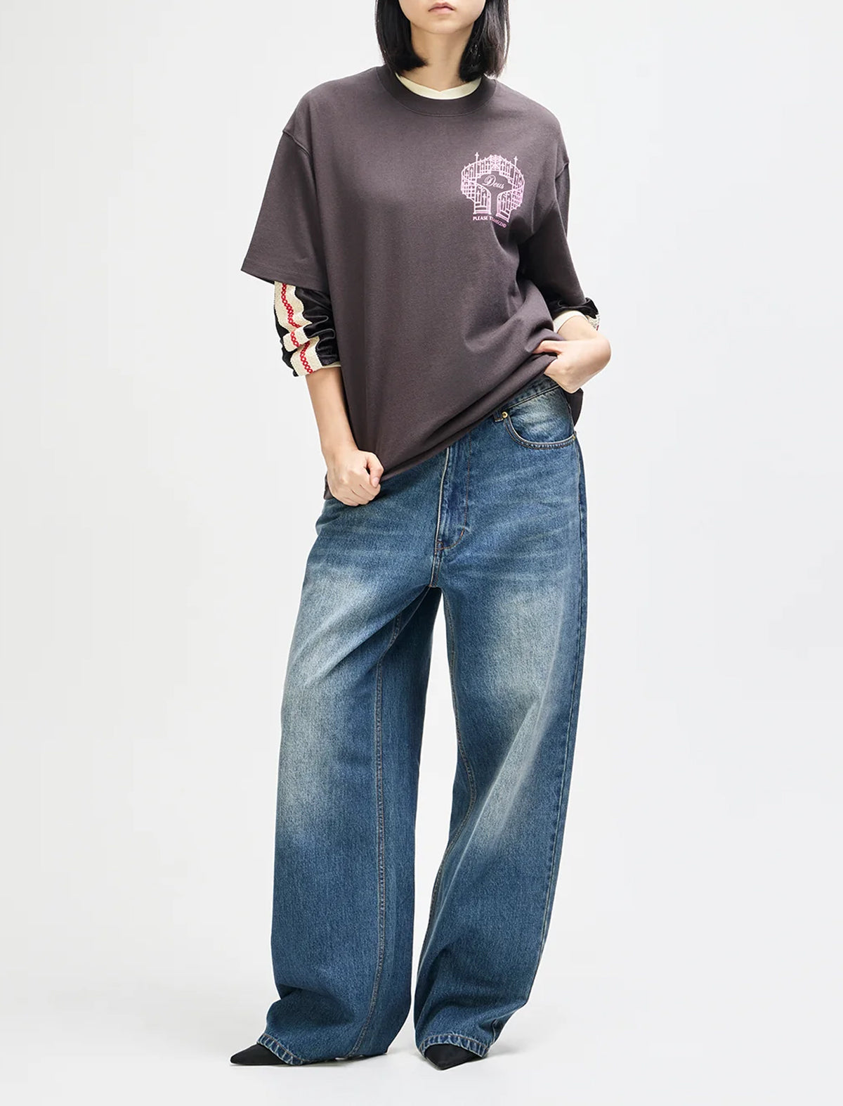 A model wearing a dark brown oversized t-shirt with a pink graphic on the chest and rolled-up sleeves, paired with wide-leg blue jeans, against a plain light gray background.