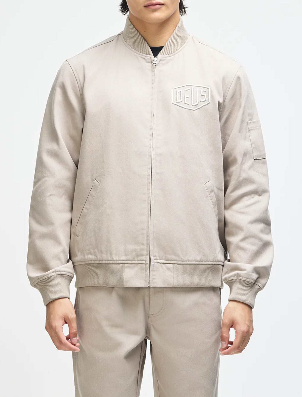 A model wearing a light beige Delius bomber jacket with a matching pair of trousers, shown from the neck down against a plain white studio background.