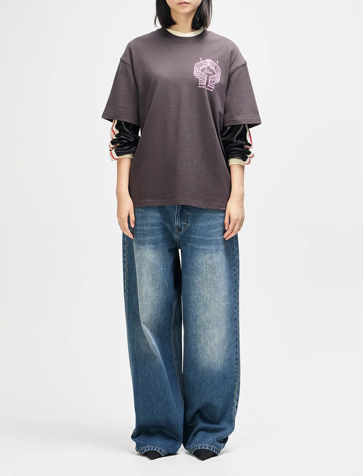 A model wearing an oversized dark brown t-shirt with a pink graphic on the chest, layered over black long-sleeve sleeves, paired with wide-leg blue jeans, against a plain white studio background.