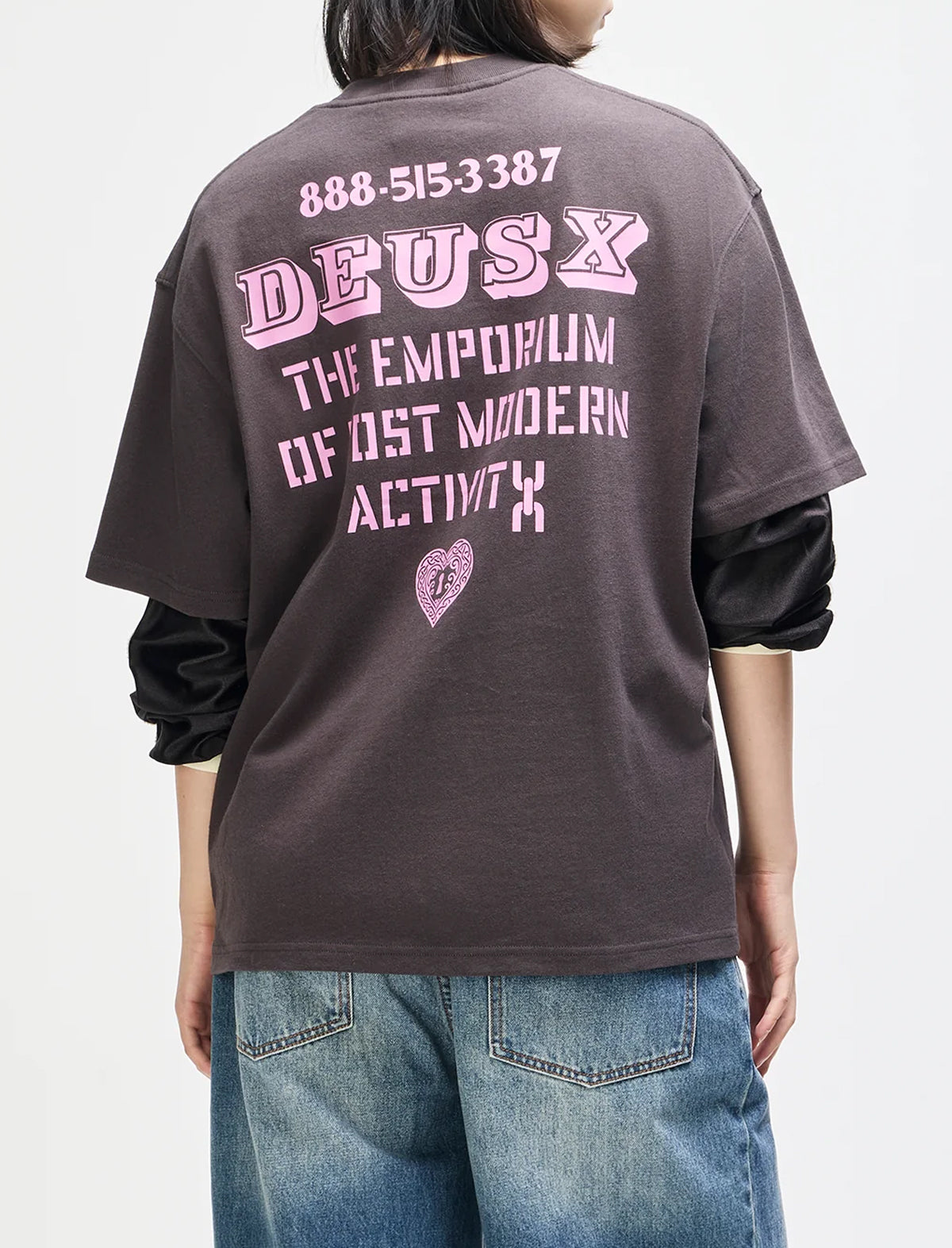 A person shown from the back wearing an oversized dark brown graphic T-shirt with pink text reading 'DEUS X' and additional slogan text, layered over a black long-sleeve shirt and blue jeans against a light gray studio background.