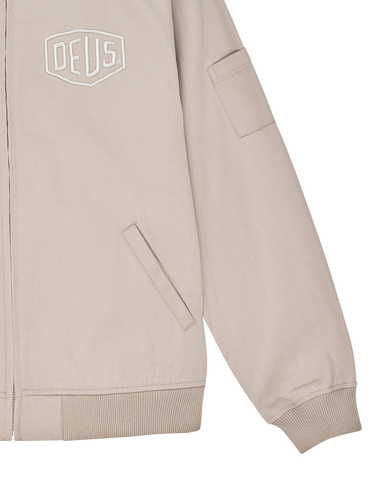 Close-up of a light beige jacket with a white outlined DEUS logo on the left chest, ribbed cuffs and hem, and a sleeve pocket, shown on a white background.