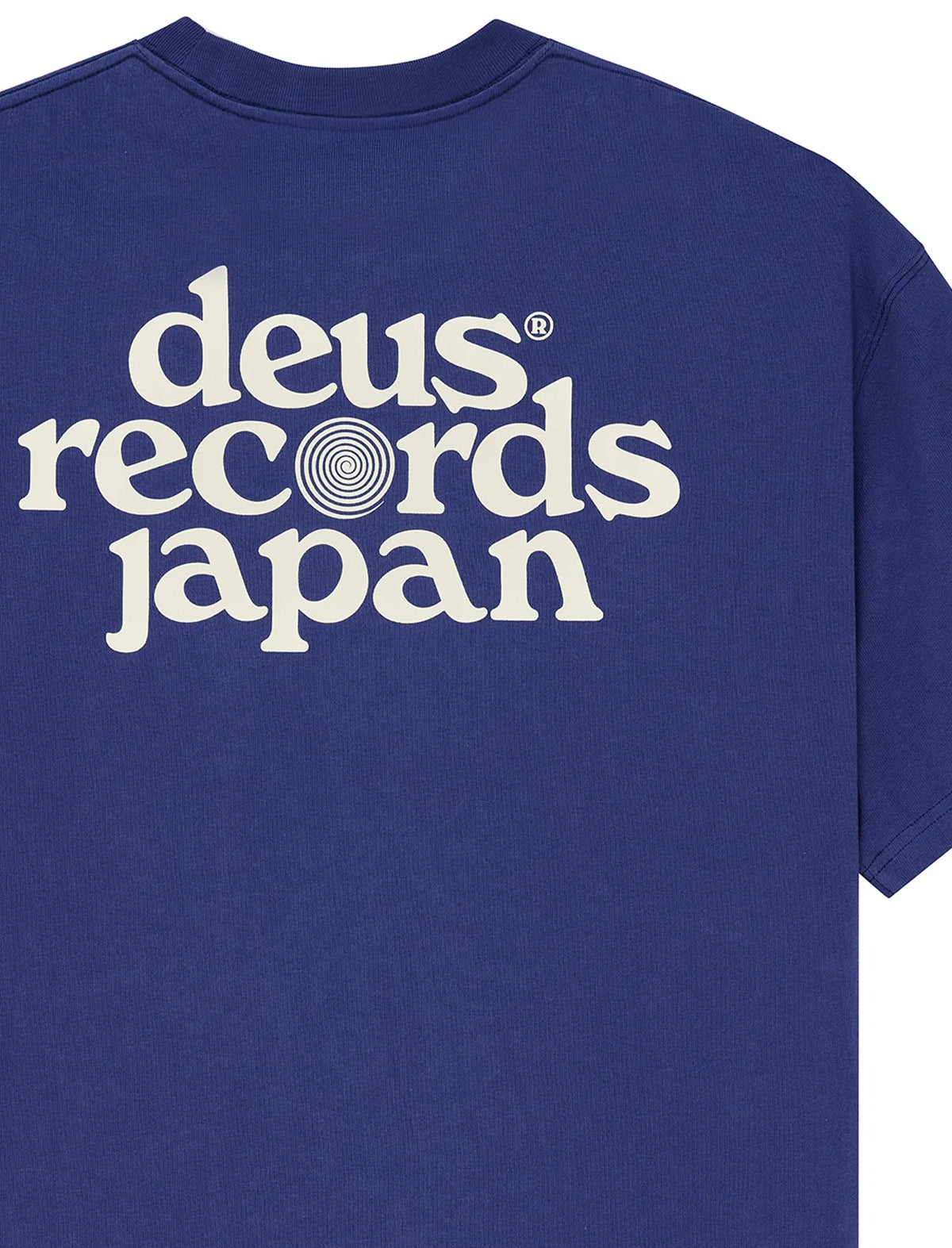 A close-up of a royal blue t-shirt showing the back, which features a large white retro-style graphic reading 'deus records japan' with a spiral design inside the letter 'o'.