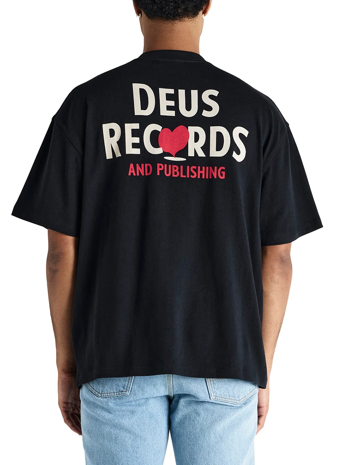 A person shown from the back wearing a black t-shirt with a large graphic print on the back reading 'DEUS RECORDS AND PUBLISHING' in white and red, paired with light blue jeans against a plain white background.