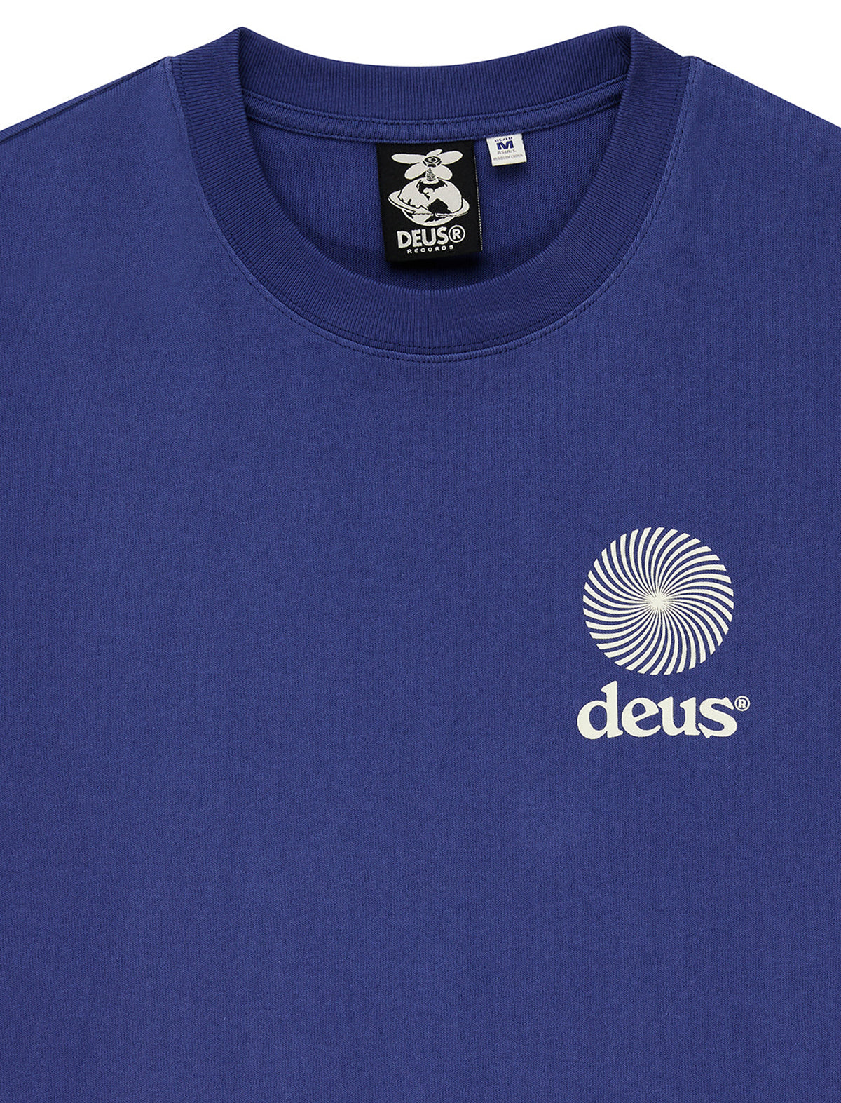 A close-up of a royal blue crewneck t-shirt featuring a white circular spiral logo and the word 'deus' printed on the left chest. The inner collar label shows the Deus brand logo.