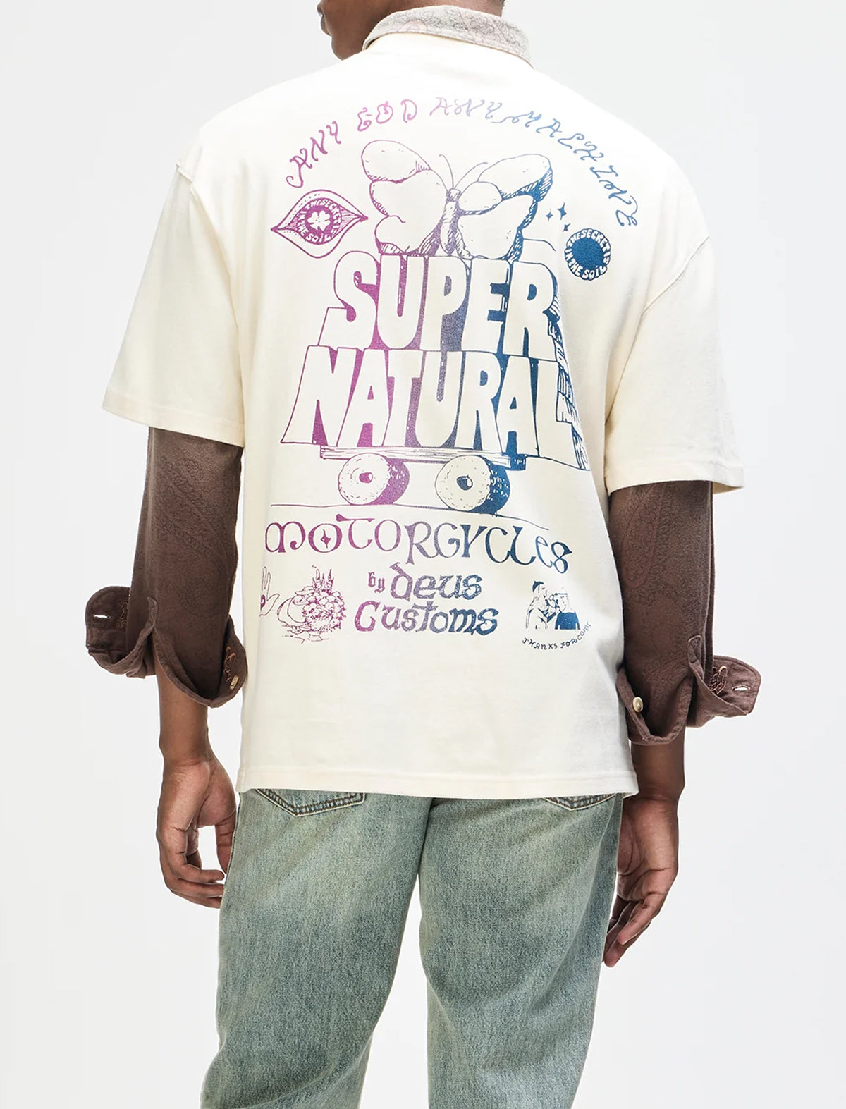 A person shown from the back wearing a cream-colored short-sleeve graphic T-shirt with a large multicolor print on the back, paired with light-wash jeans. The shirt features text and illustrations including 'SUPER NATURAL' and 'MOTORCYCLES by Deus Customs'.