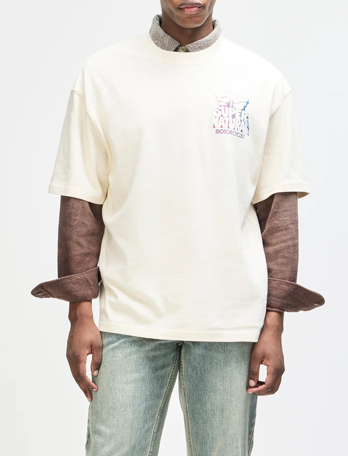 A model wearing a cream-colored short-sleeve t-shirt with a brown textured long-sleeve layer underneath, styled with light-wash jeans against a plain white background.