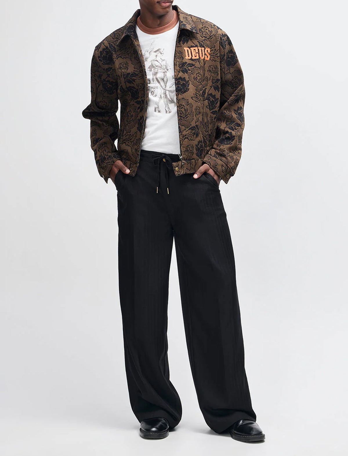 Person wearing a patterned jacket, white shirt with 'DEUS' logo, and black pants on a white background