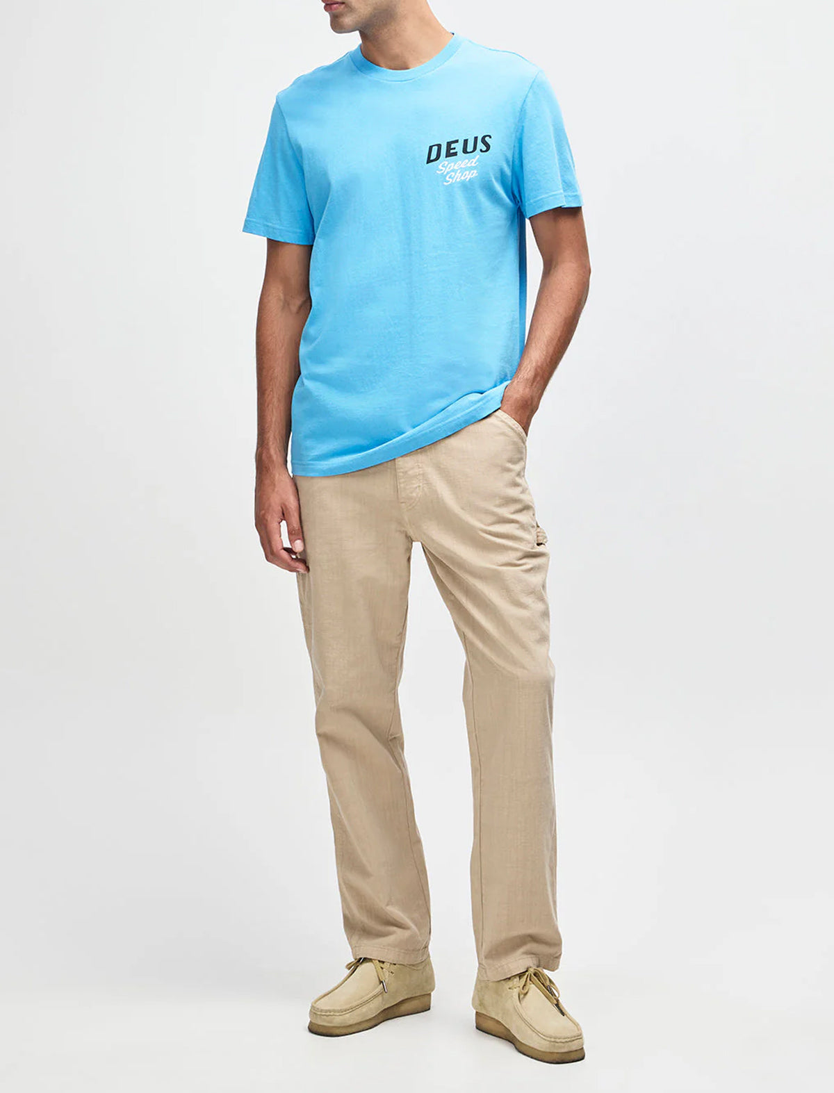 Person wearing a light blue t-shirt with 'DEUS' logo and beige pants on a white background