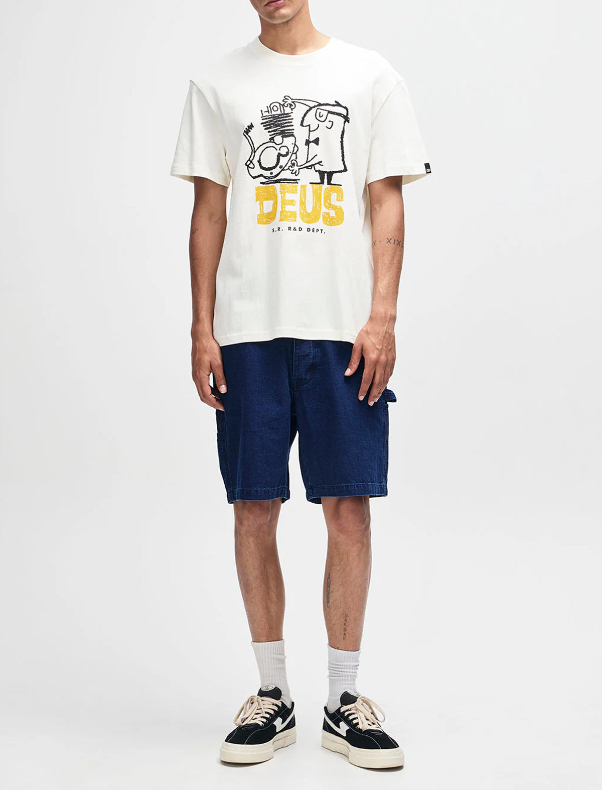 Person wearing a white t-shirt with a logo and text, navy shorts, and black sneakers on a white background