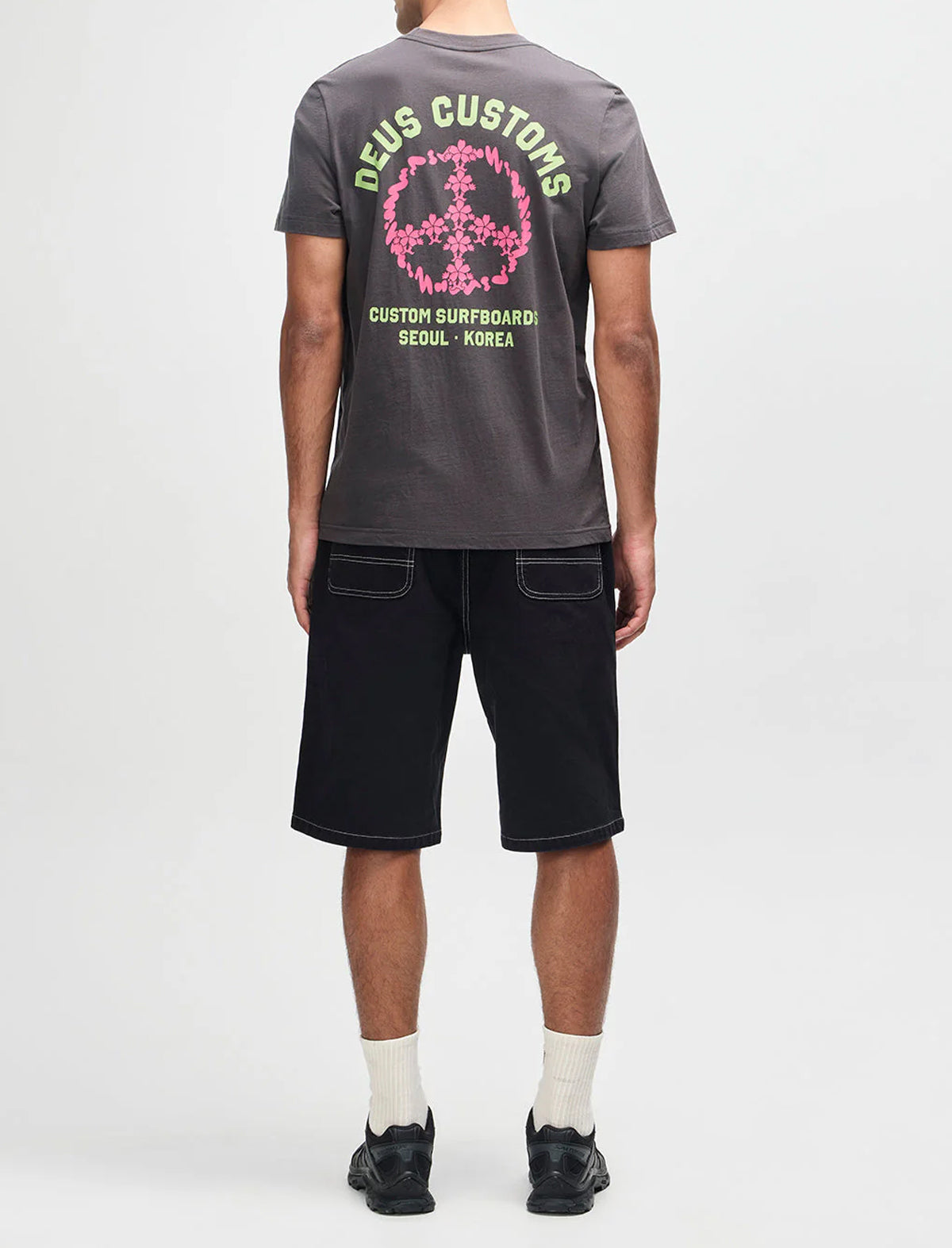 Person wearing a gray 'Deus Customs' t-shirt with a pink and green graphic on the back, standing against a white background.